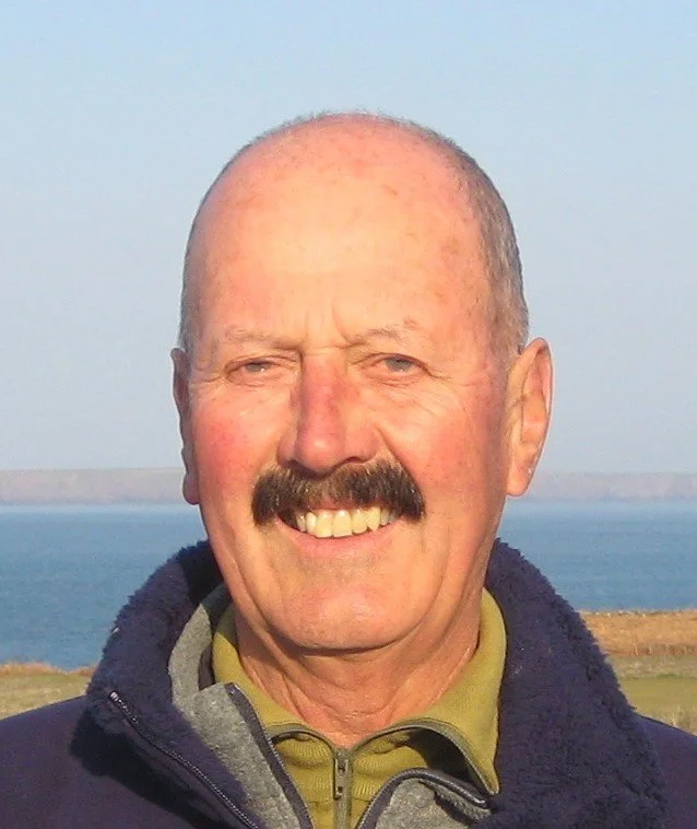Close-up of a smiling middle-aged man with a mustache, outdoors near water, wearing a dark jacket and a green shirt.