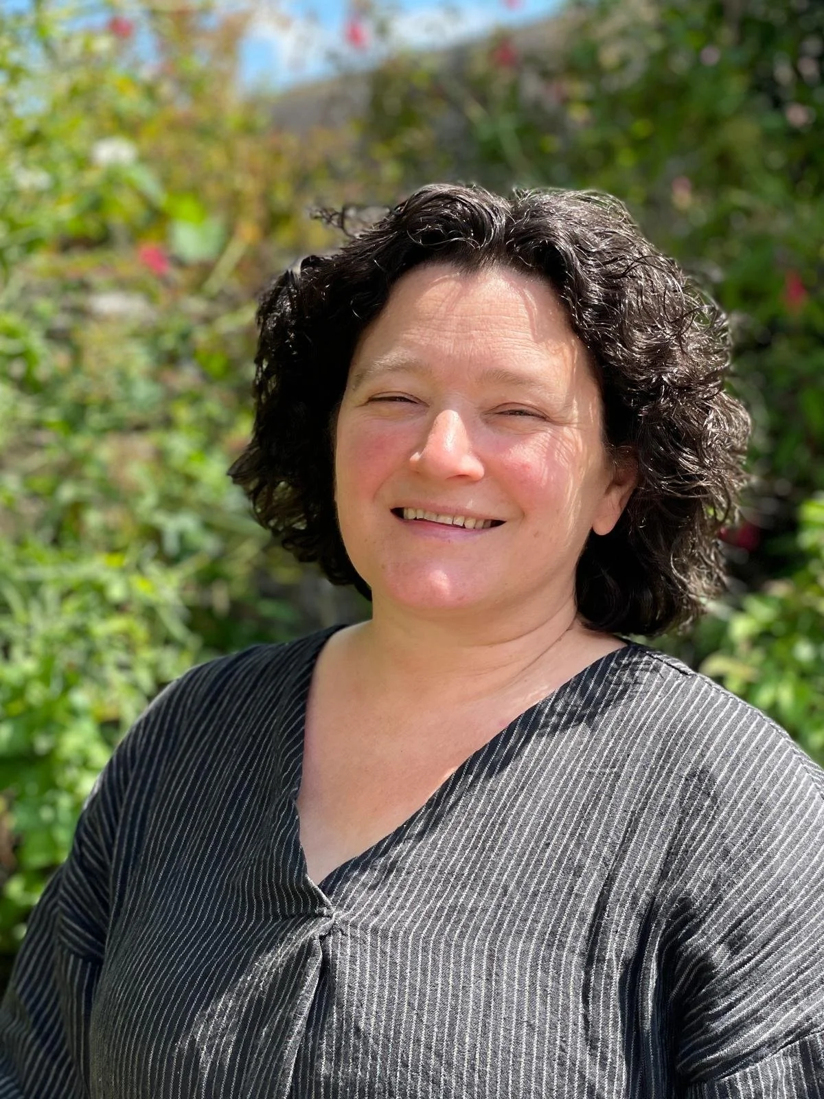 Smiling woman with curly dark hair outdoors, greenery in the background.