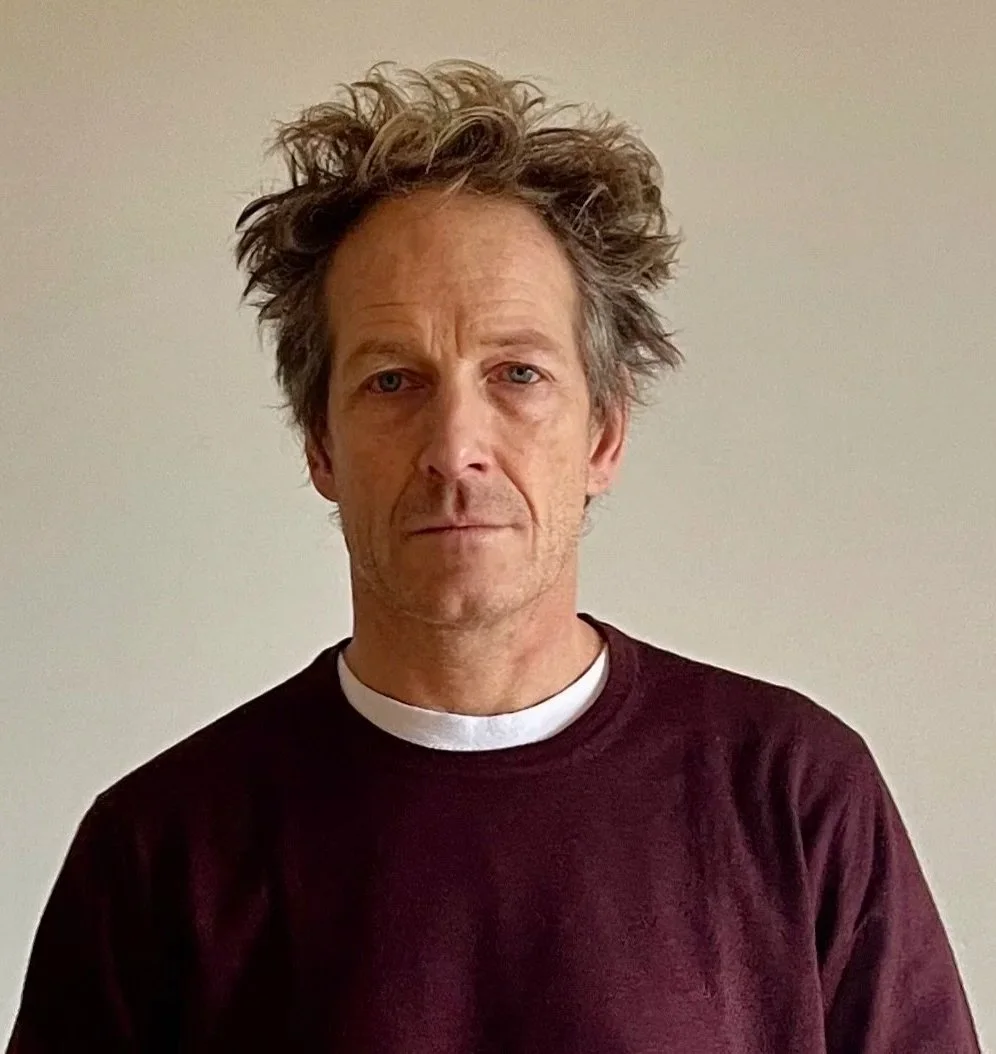 A middle-aged man with messy, unkempt hair, wearing a dark maroon sweatshirt over a white shirt, standing against a plain light-colored background.