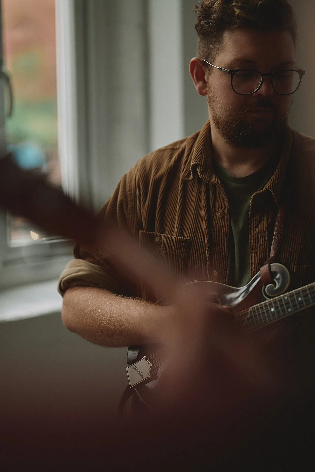 A man with glasses and a beard playing a mandolin indoors near a window, with natural light illuminating his brown jacket and green shirt.