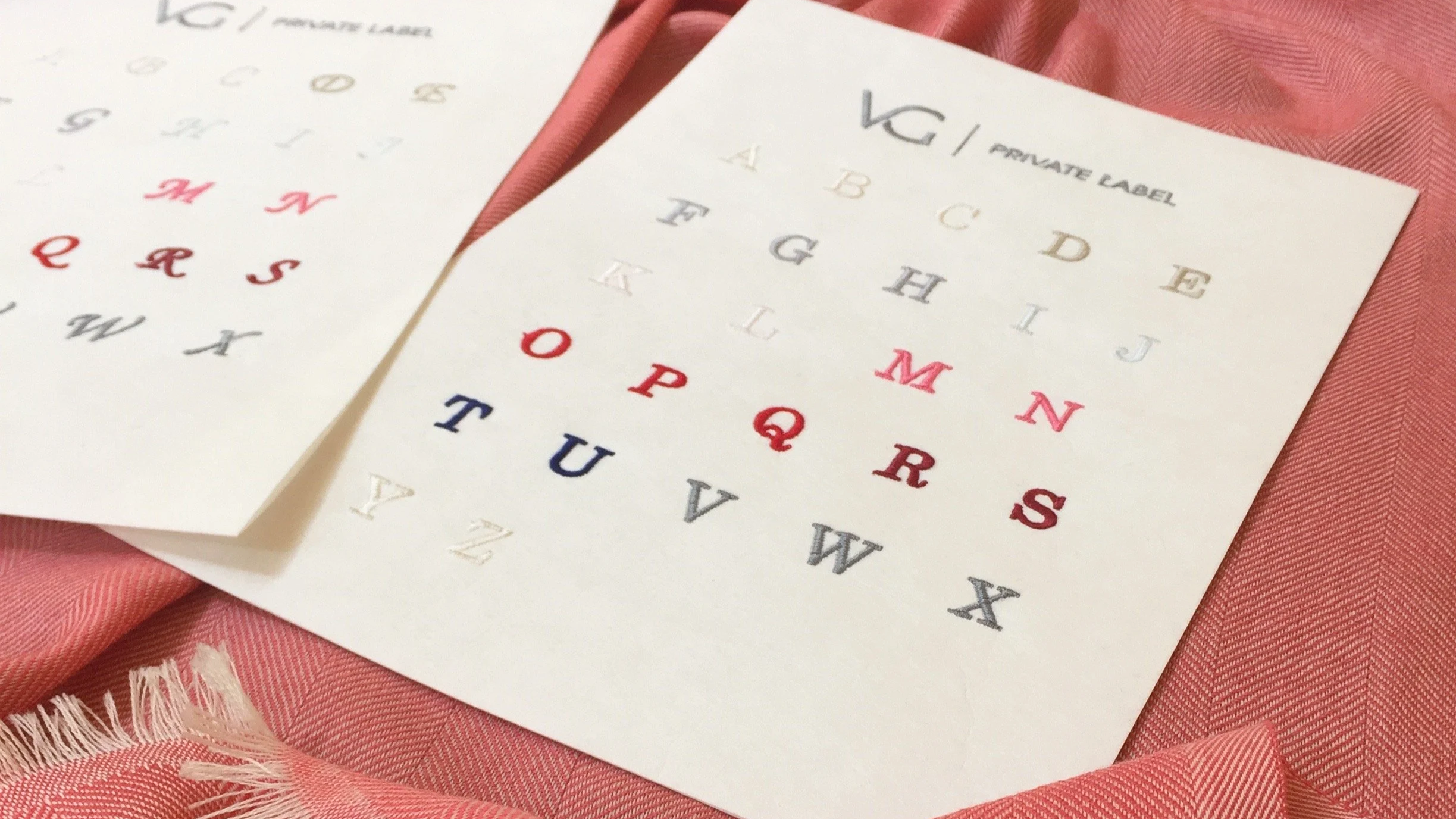 Deux feuilles avec des alphabets en différentes couleurs et styles de lettres, posées sur un tissu rose avec bord frangé.