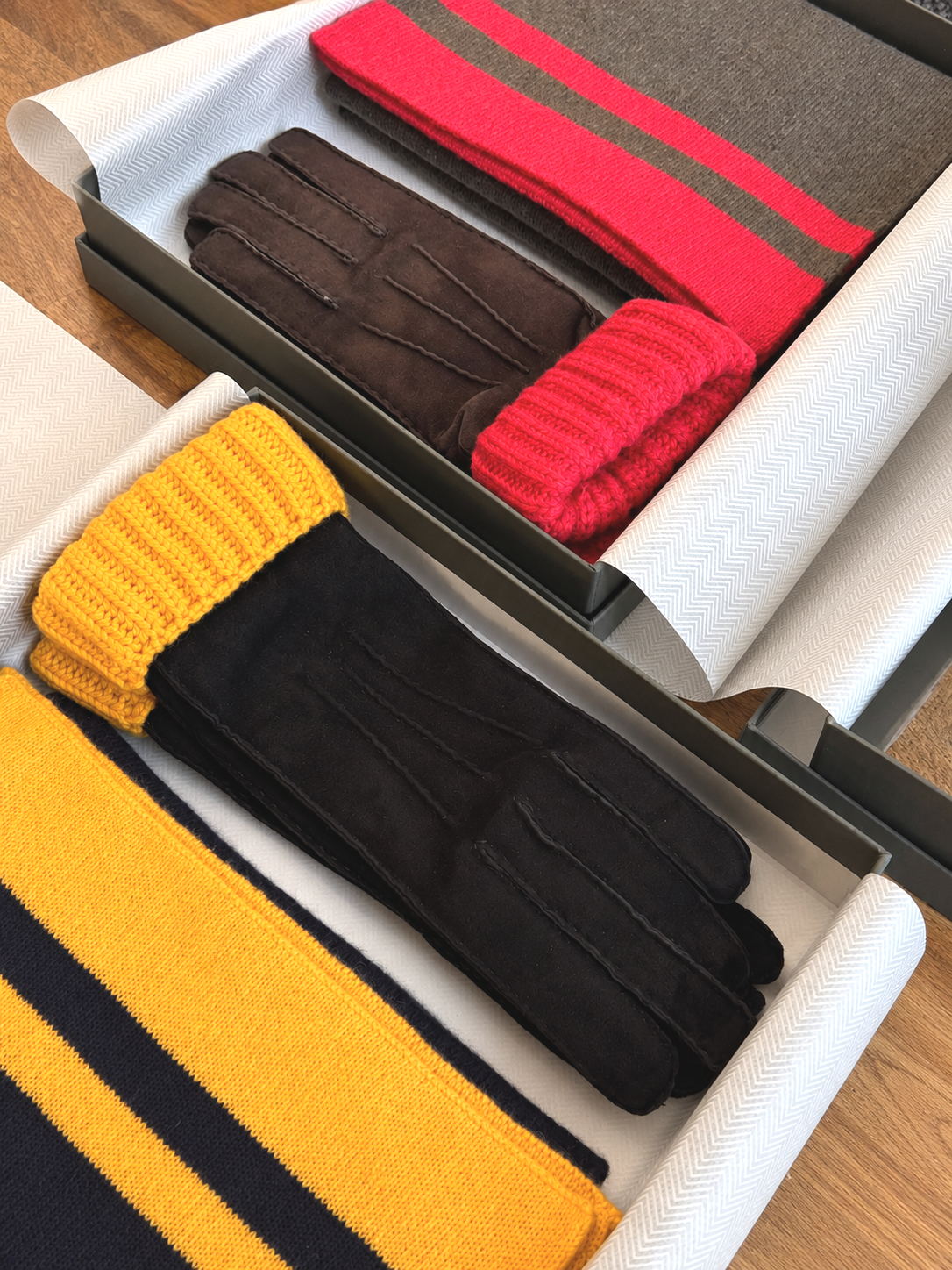 Deux boîtes contenant des accessoires en laine ou en tissu, notamment des gants noirs, des écharpes rouges et jaunes, et des bonnets rouges, noirs, et gris avec une rayure rouge. La scène est organisée sur une surface en bois.