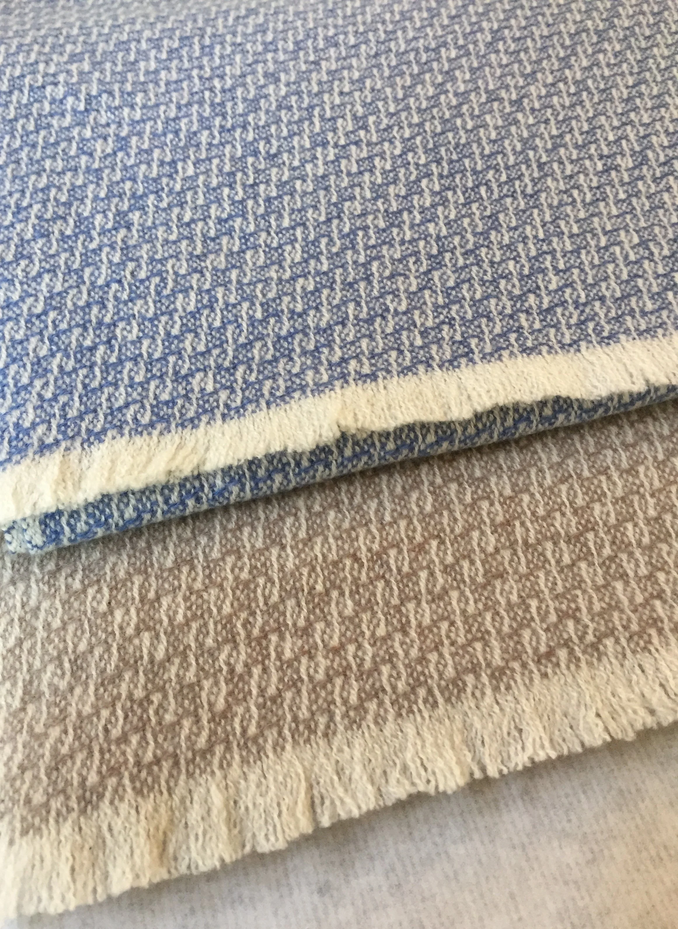 Une grande serviette en tissu blanc et une autre en tissu bleu avec un motif en forme de V ou chevrons. La serviette blanche est pliée et repose en dessous de la serviette bleue. Les deux serviettes sont posées sur un tapis beige.