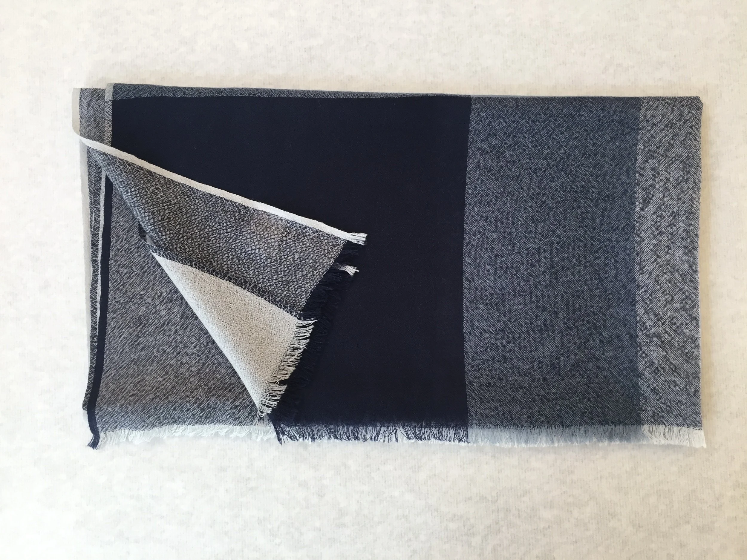 Un tapis ou une couverture en tissu gris foncé avec des franges aux extrémités, posée sur une surface claire.