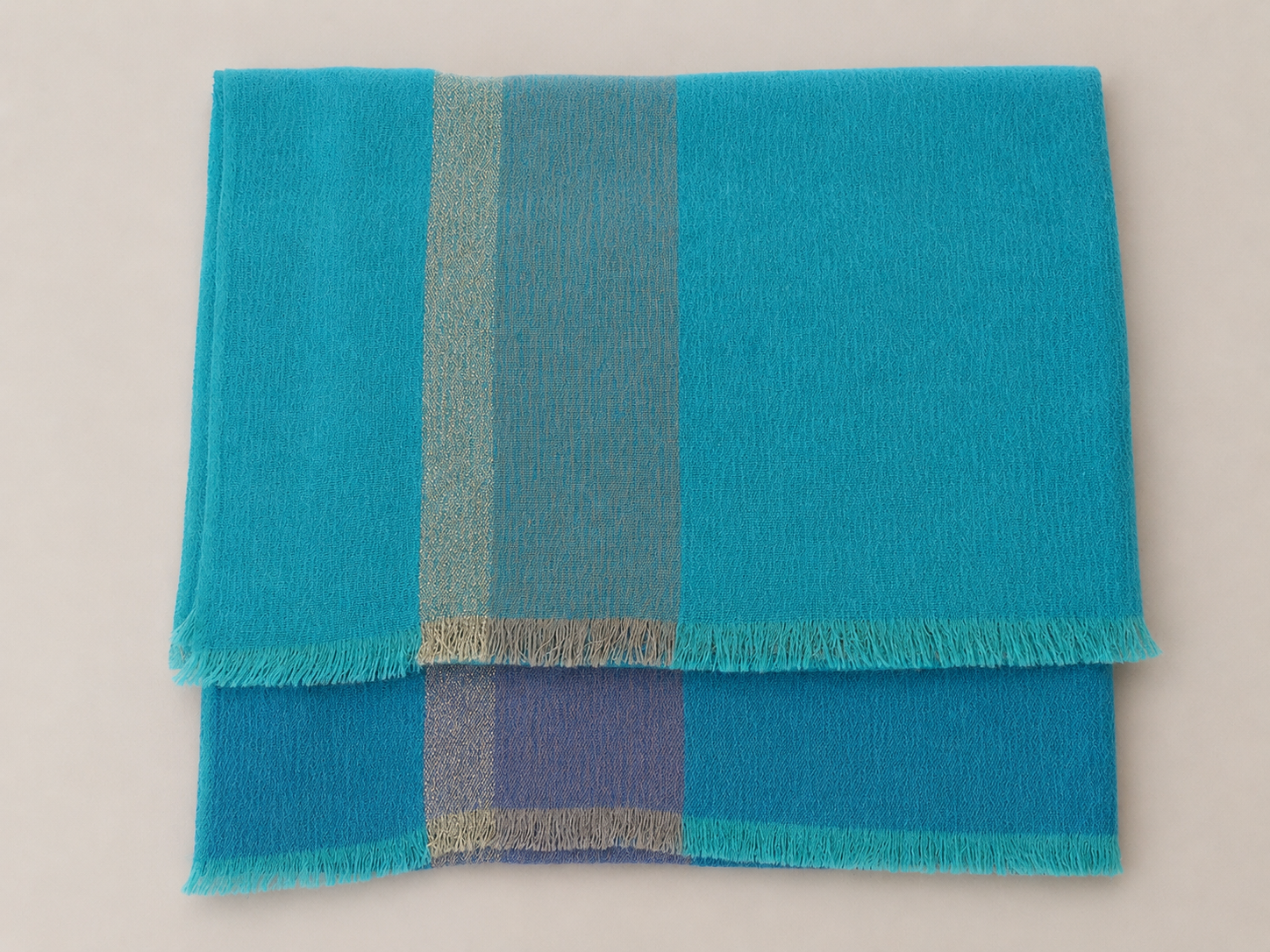 Deux serviettes de couleur turquoise avec des bandes argentées et bleues, posées l'une sur l'autre sur un fond blanc.