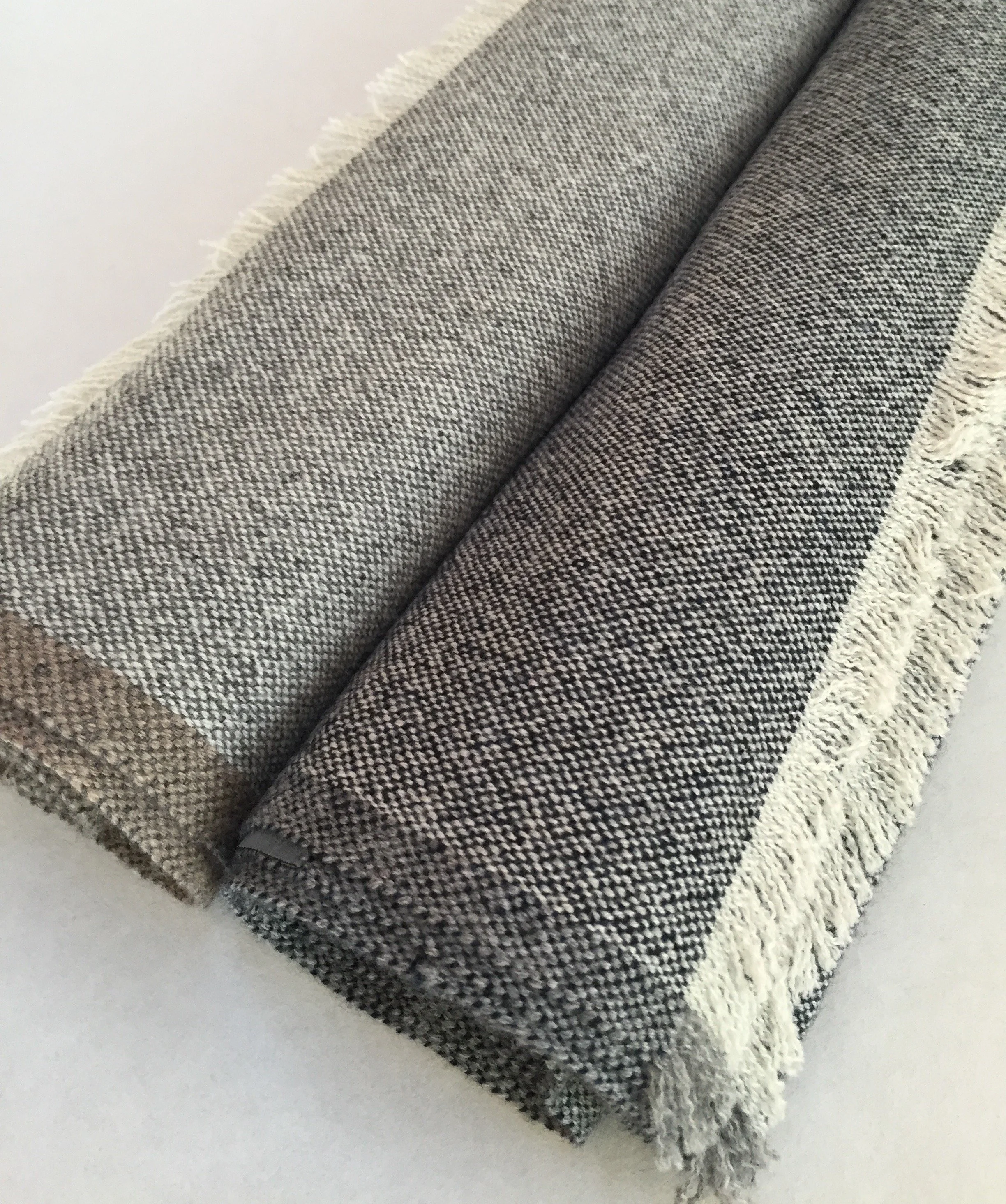 Un tissu tissé gris avec des bordures à franges blanches, posé sur une surface blanche.