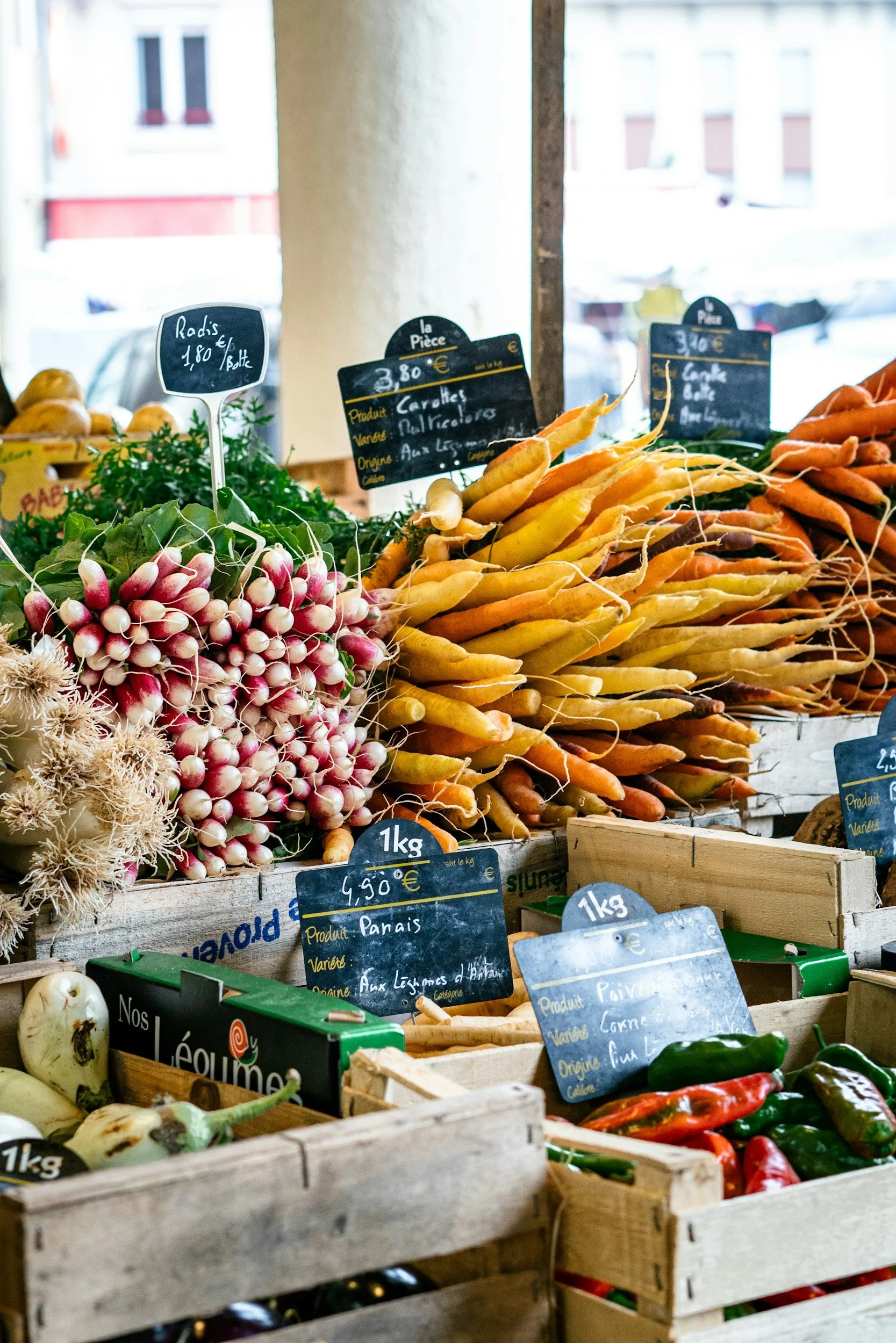 Légumes frais dans un marché, notamment des carottes jaunes et oranges, des radis roses et blancs, et des piments verts et rouges, avec des pancartes indiquant leur prix et leur origine.