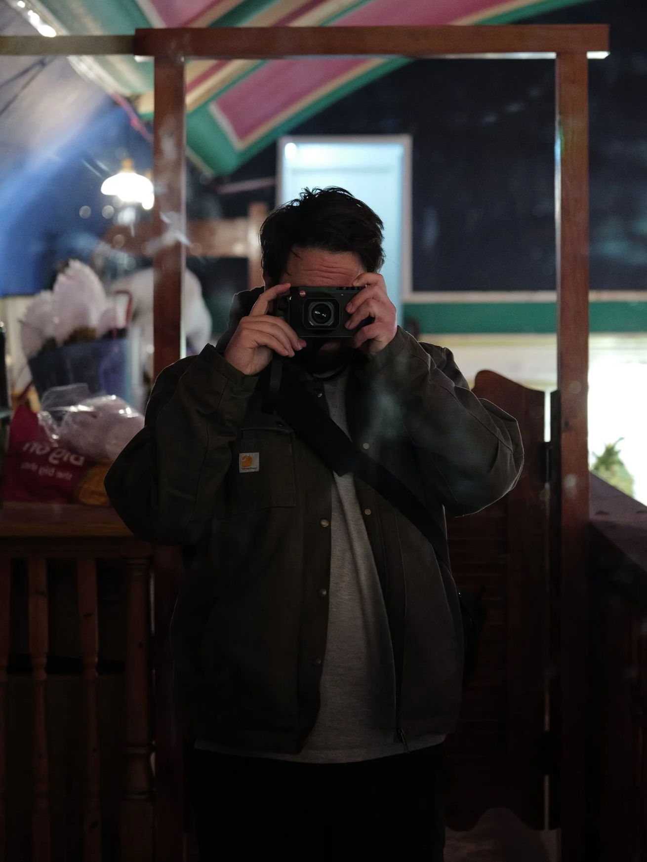 A person taking a mirror selfie indoors, holding a camera up to their face, wearing a dark jacket and gray shirt, with a colorful ceiling in the background.