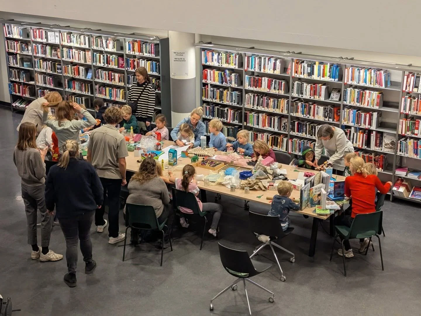 People, including children and adults, gathered around a large table in a library, engaging in arts and crafts activities, with bookshelves in the background.