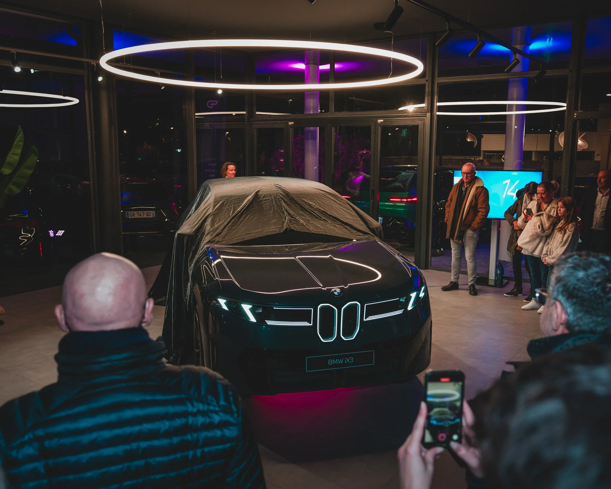 Une voiture électrique BMW iX3 dévoilée lors d'une présentation en intérieur, entourée de spectateurs et de personnes prenant des photos.