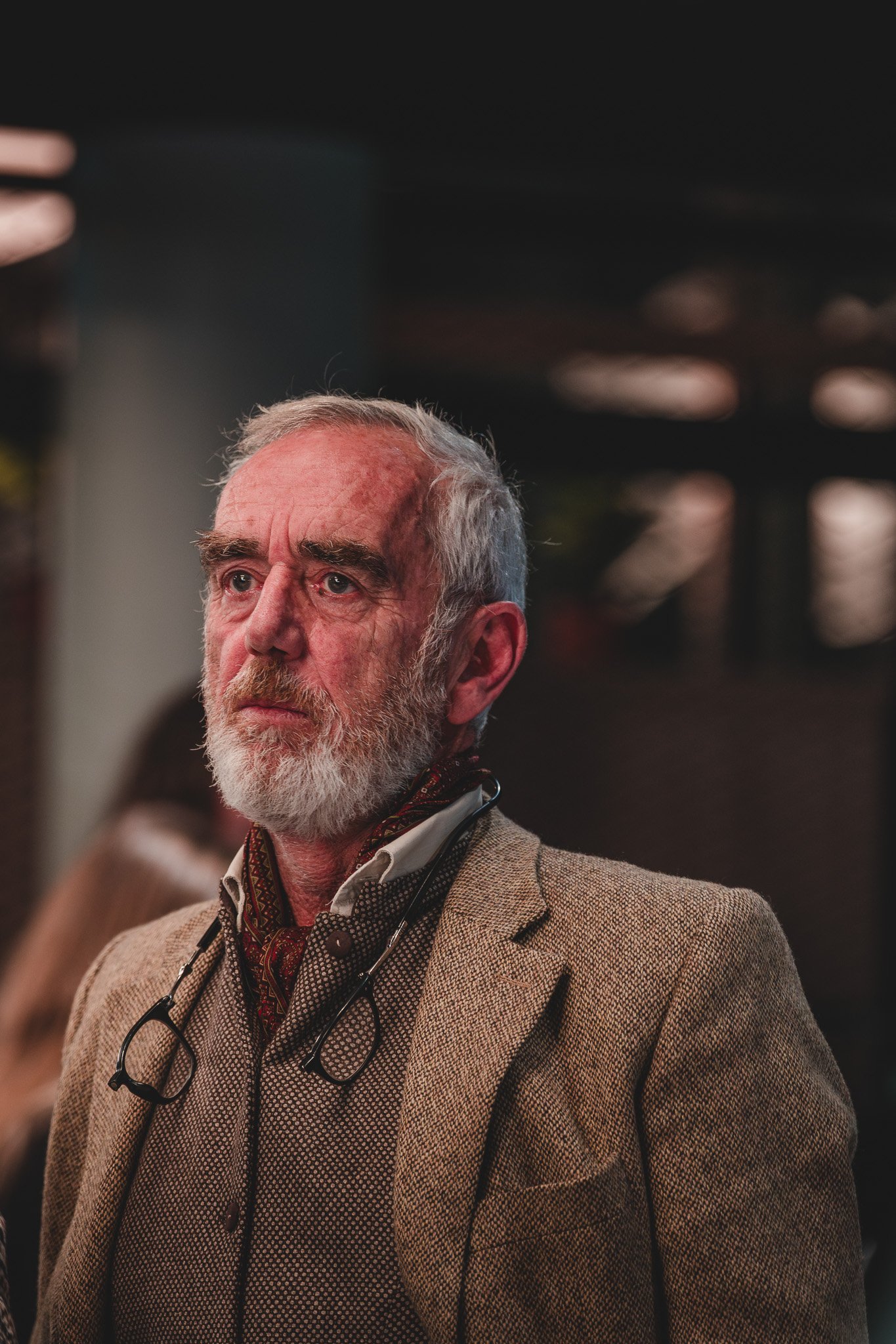 Un homme âgé avec une barbe grise et des cheveux gris, portant un costume marron avec une chemise blanche, dans un environnement intérieur.