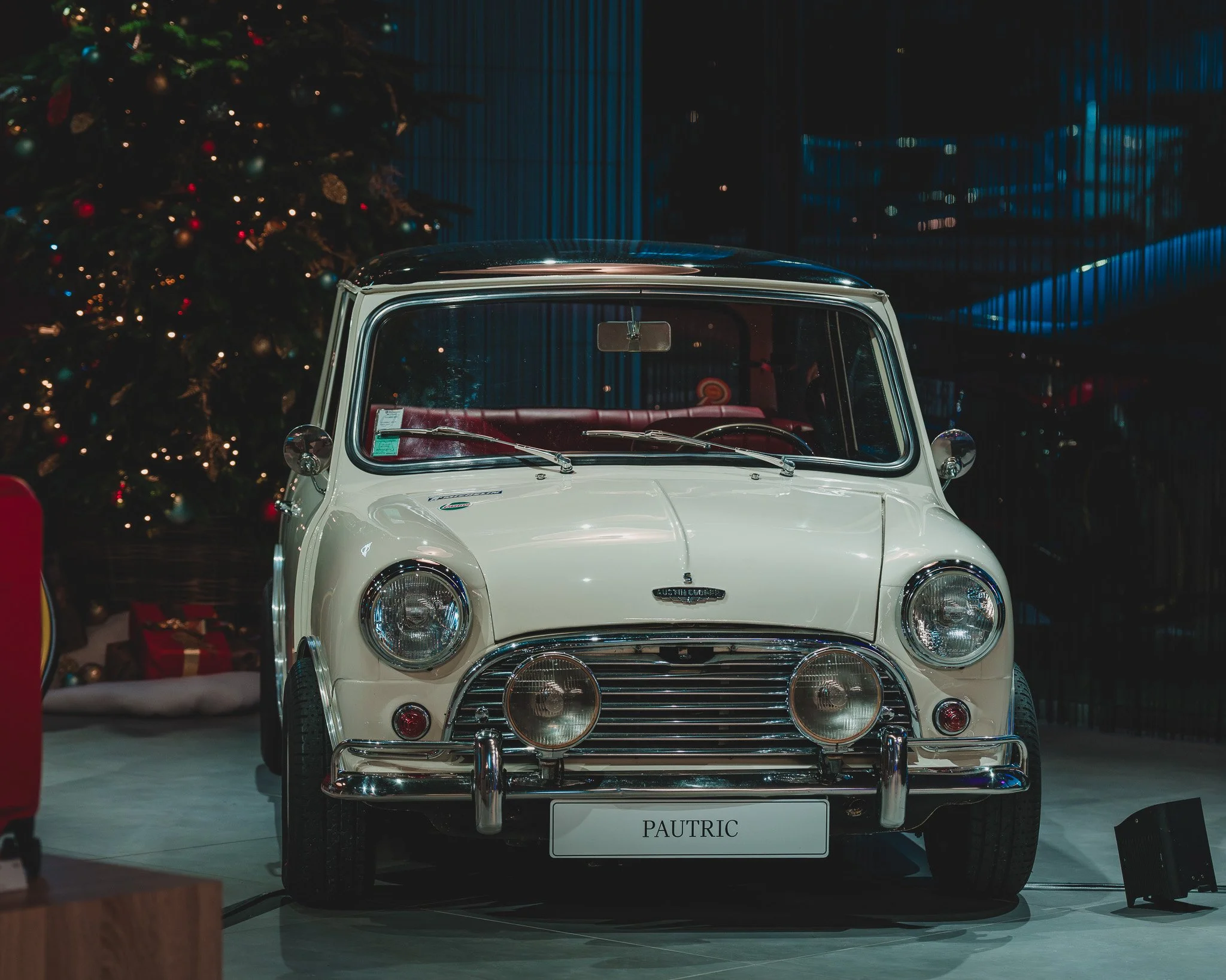 Voiture blanche de style rétro avec un pare-chocs chromé, exposée en intérieur, décorée pour Noël avec un sapin et des cadeaux en arrière-plan.