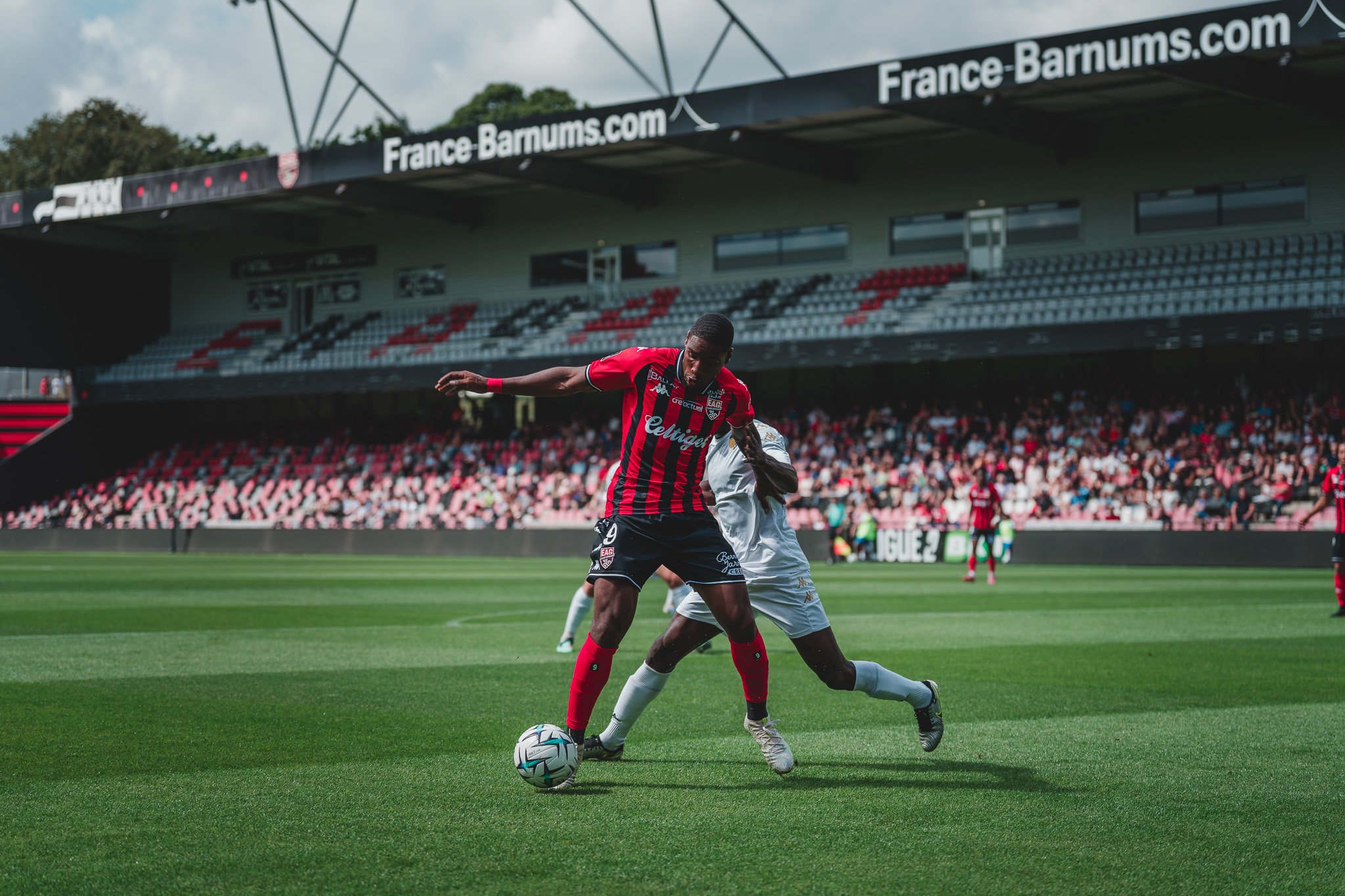 Deux joueurs de football, un en maillot rouge et noir et l'autre en blanc, en duel pour le ballon sur un terrain de football avec un stade et des spectateurs en arrière-plan.