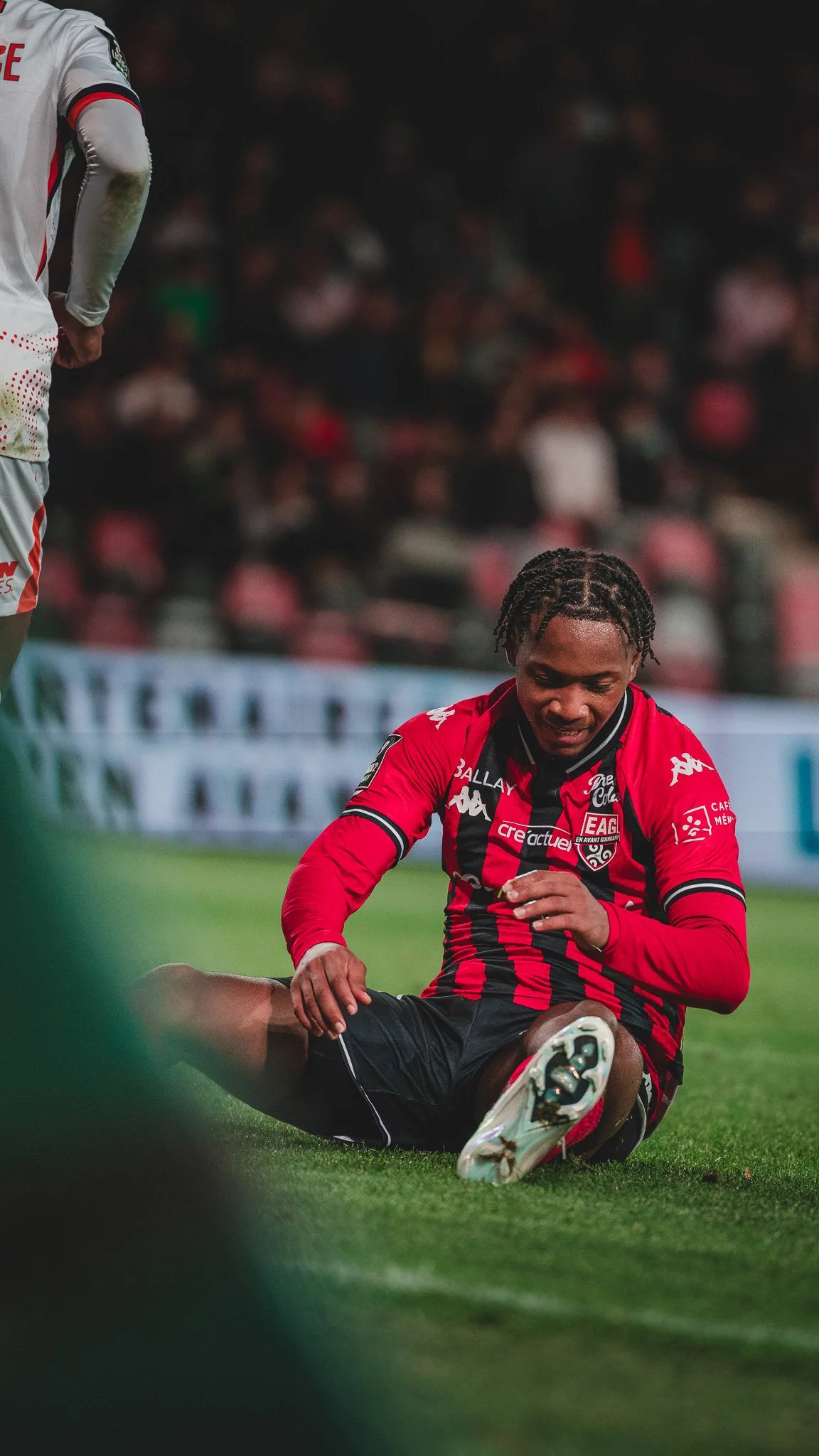 Un joueur de football en tenue rouge et noire, assis sur le sol, après avoir été blessé lors d'un match, avec des spectateurs en arrière-plan.
