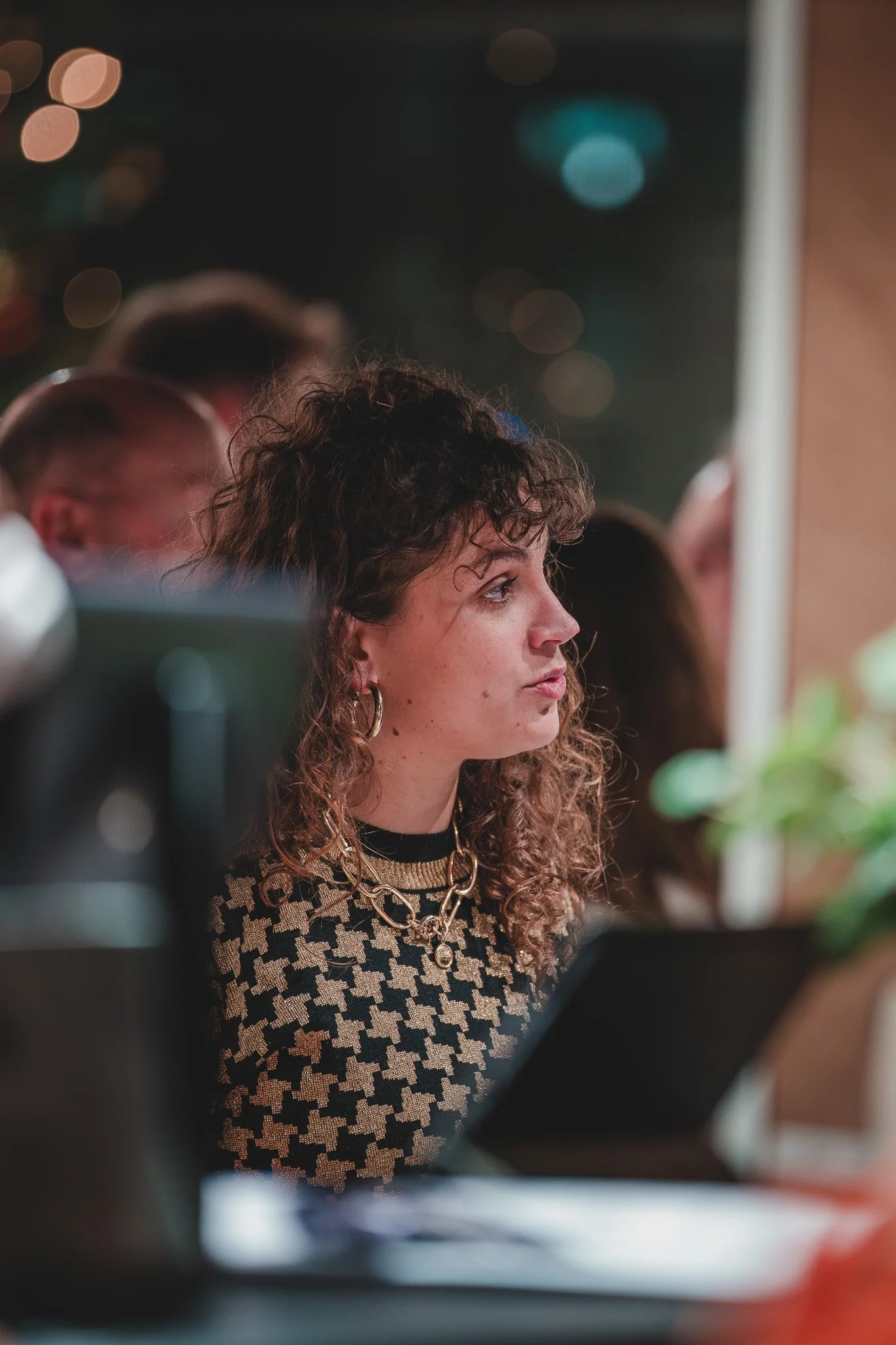 Femme assise à un bureau, en pleine concentration, portant un pull à motif pied-de-poule, des boucles d'oreilles en cercle et une chaîne en or.