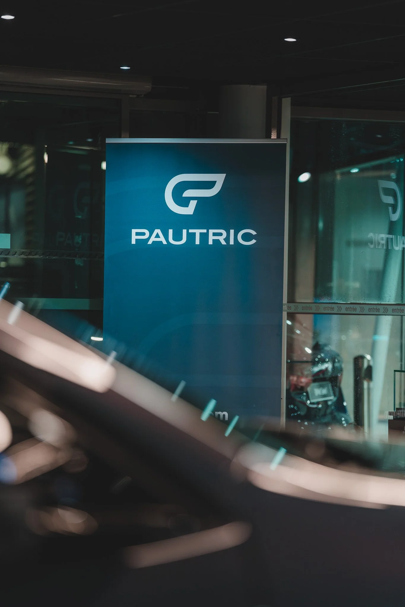 Panneau d'affichage avec le logo de Pautric devant une entrée de bâtiment, avec des voitures visibles derrière la vitre.