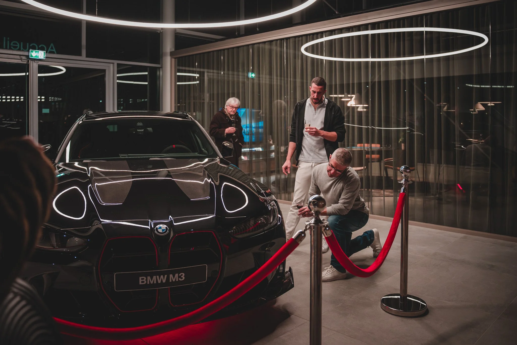 Une voiture BMW M3 noire exposée en showroom, entourée d'une barrière rouge, avec trois personnes observant et discutant autour, dans un environnement moderne avec éclairage circulaire au plafond.