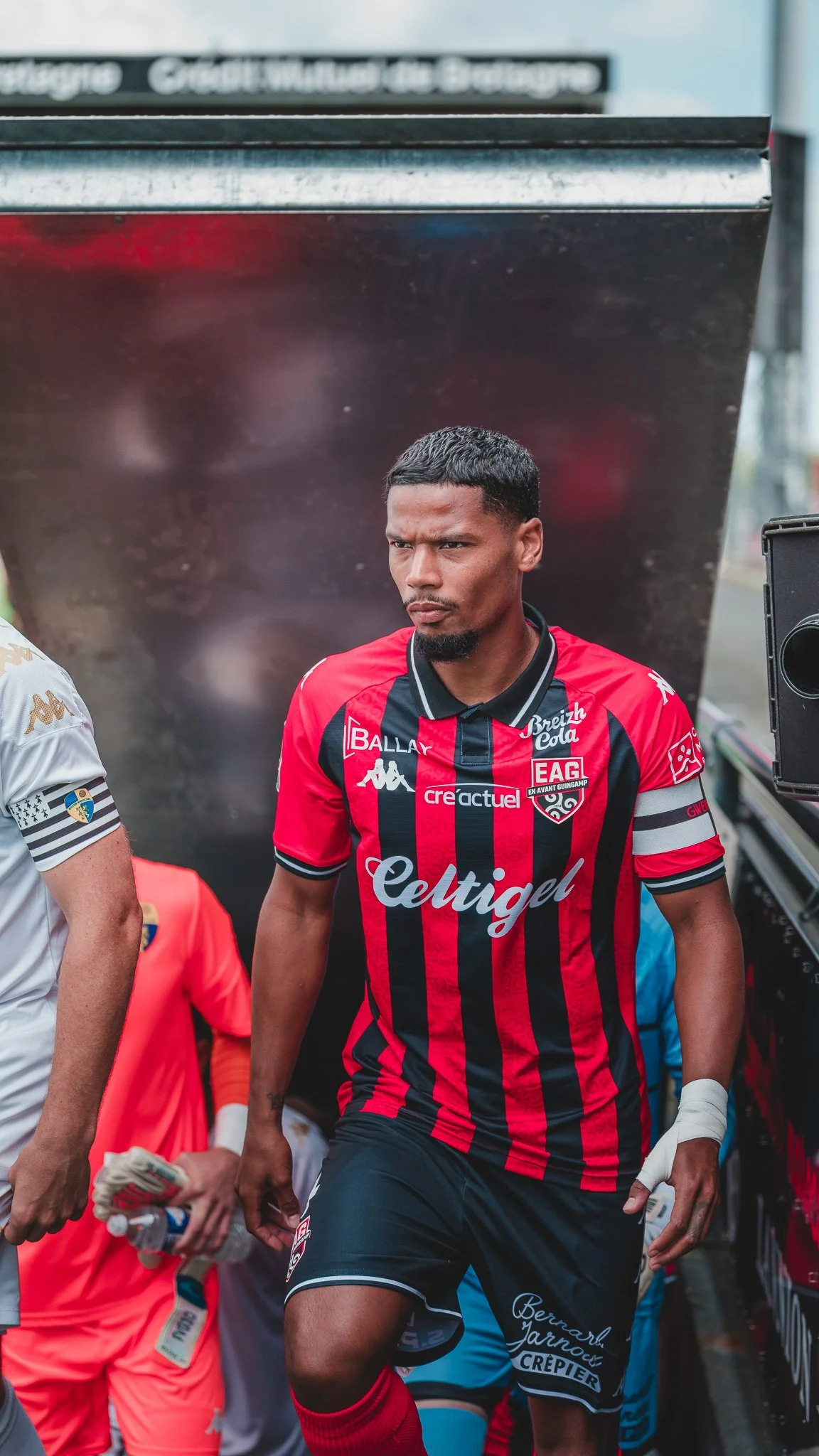 Un joueur de football en tenue rouge et noire avec le logo 'Celtigel' sur la poitrine, marche dans un tunnel de stade.