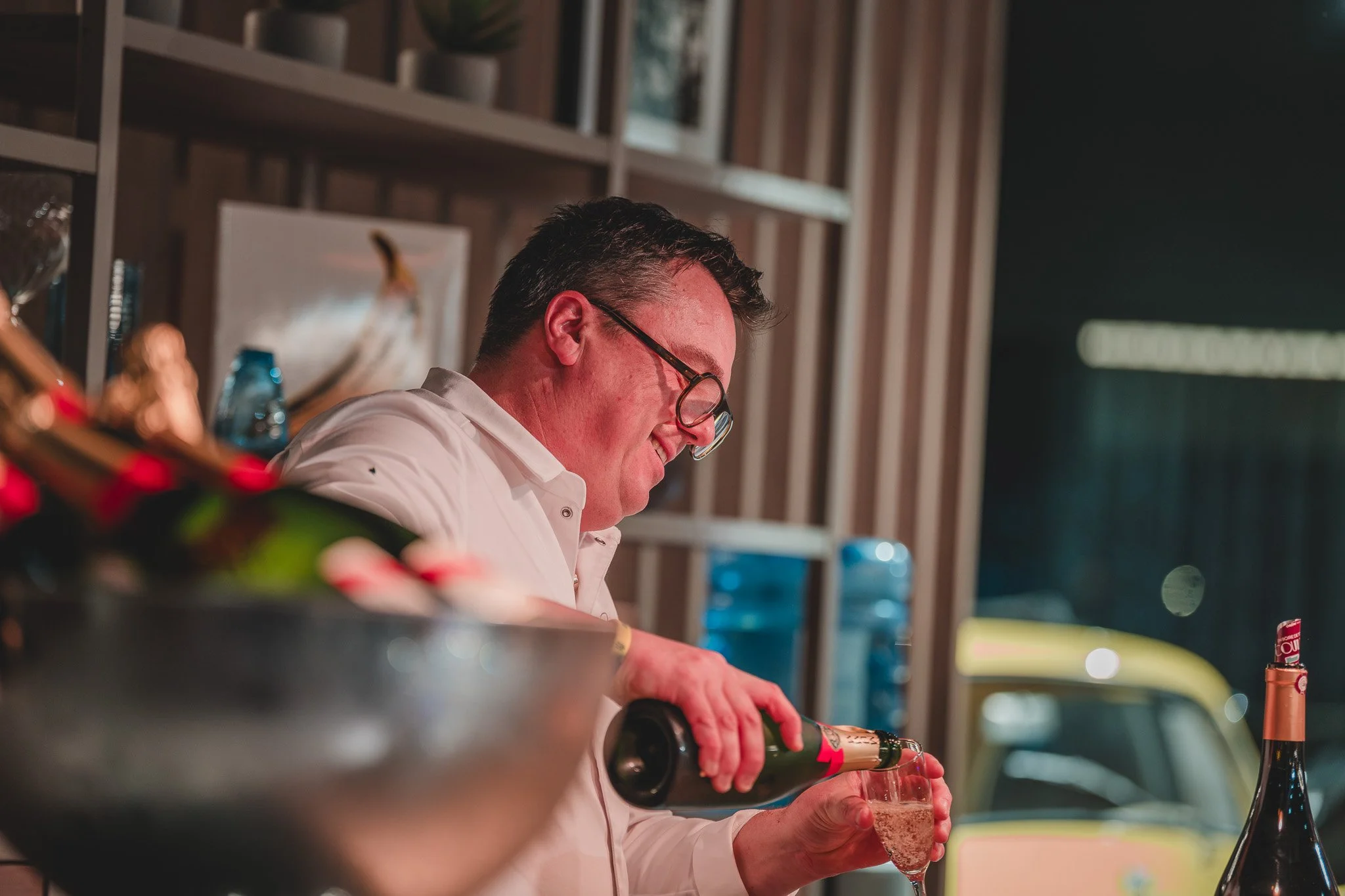 Un homme souriant verse du champagne dans une flûte, derrière un bar, en intérieur, à côté d'une voiture jaune visible par la fenêtre.