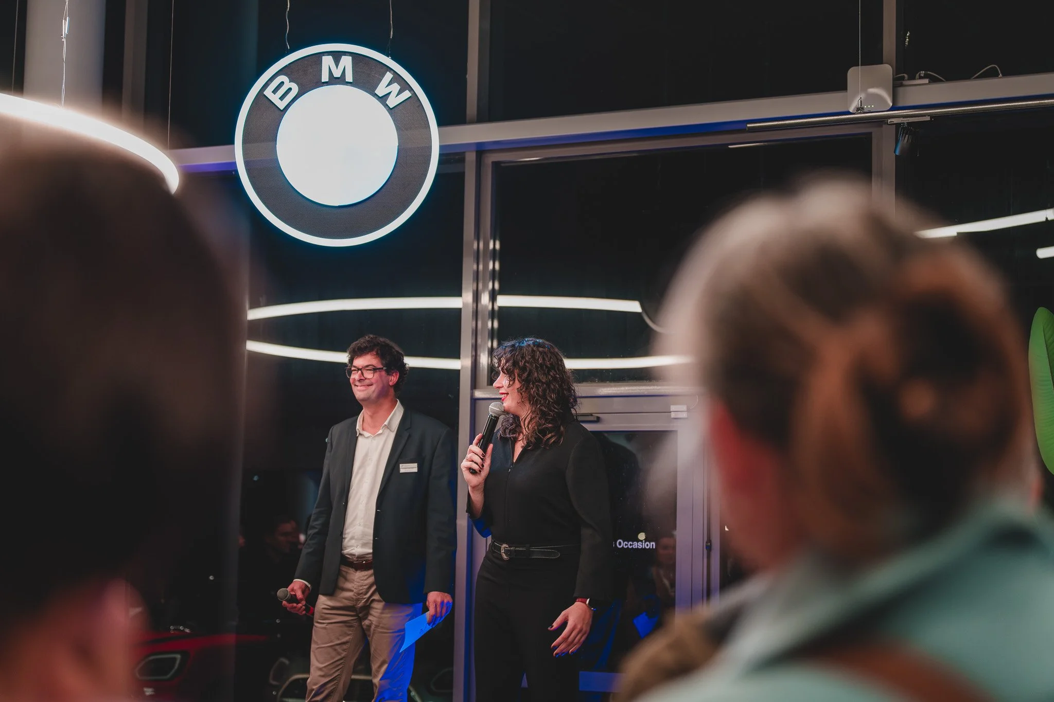 Deux personnes parlant lors d'un événement dans un centre BMW, un homme en costume et une femme tenant un microphone, entourés de plusieurs spectateurs, avec un logo BMW visible en arrière-plan.