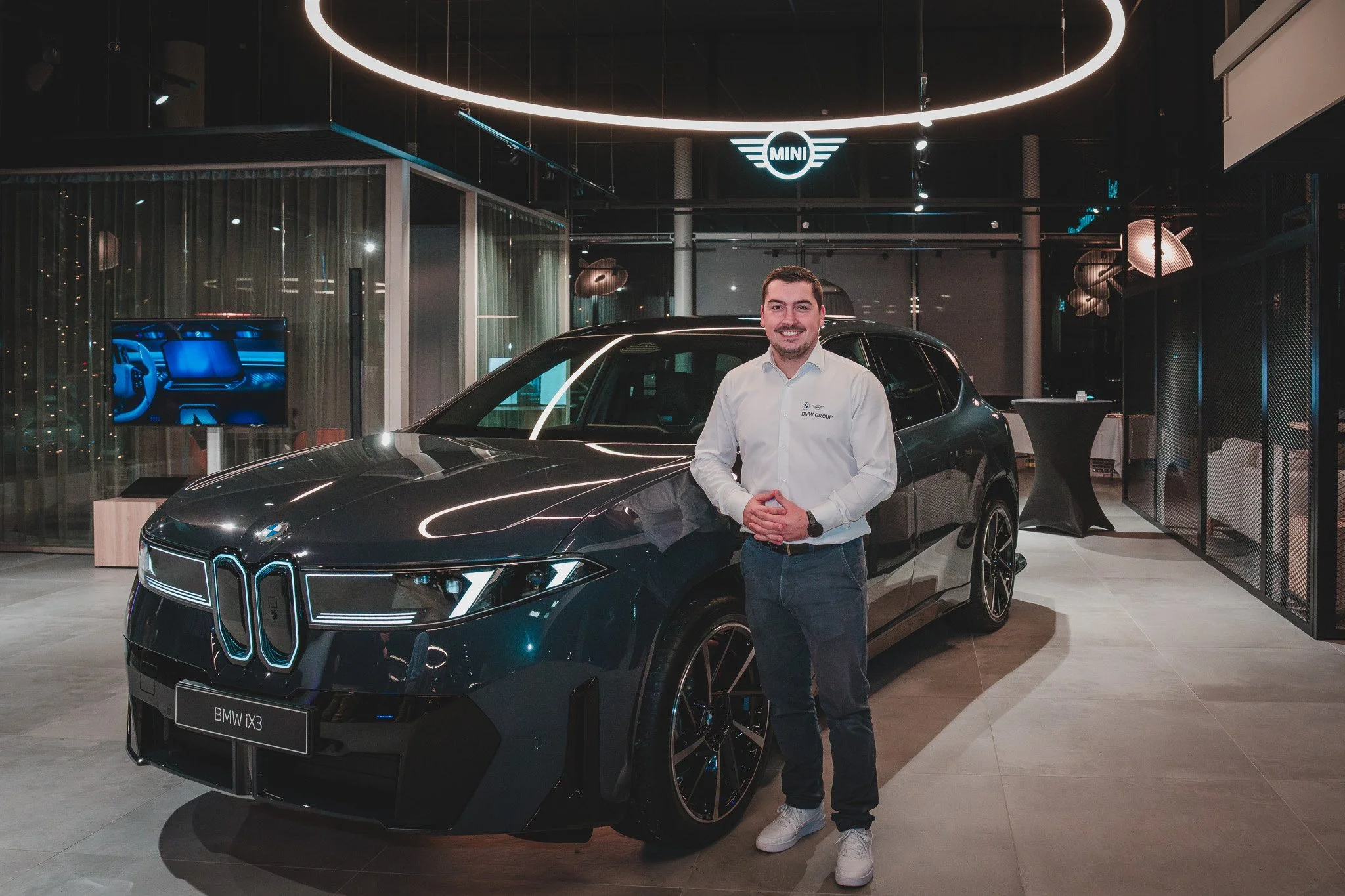 Un homme souriant posant à côté d'une voiture noire dans un showroom automobile, avec des panneaux lumineux, un logo MINI suspendu au plafond et un écran numérique à l'arrière.
