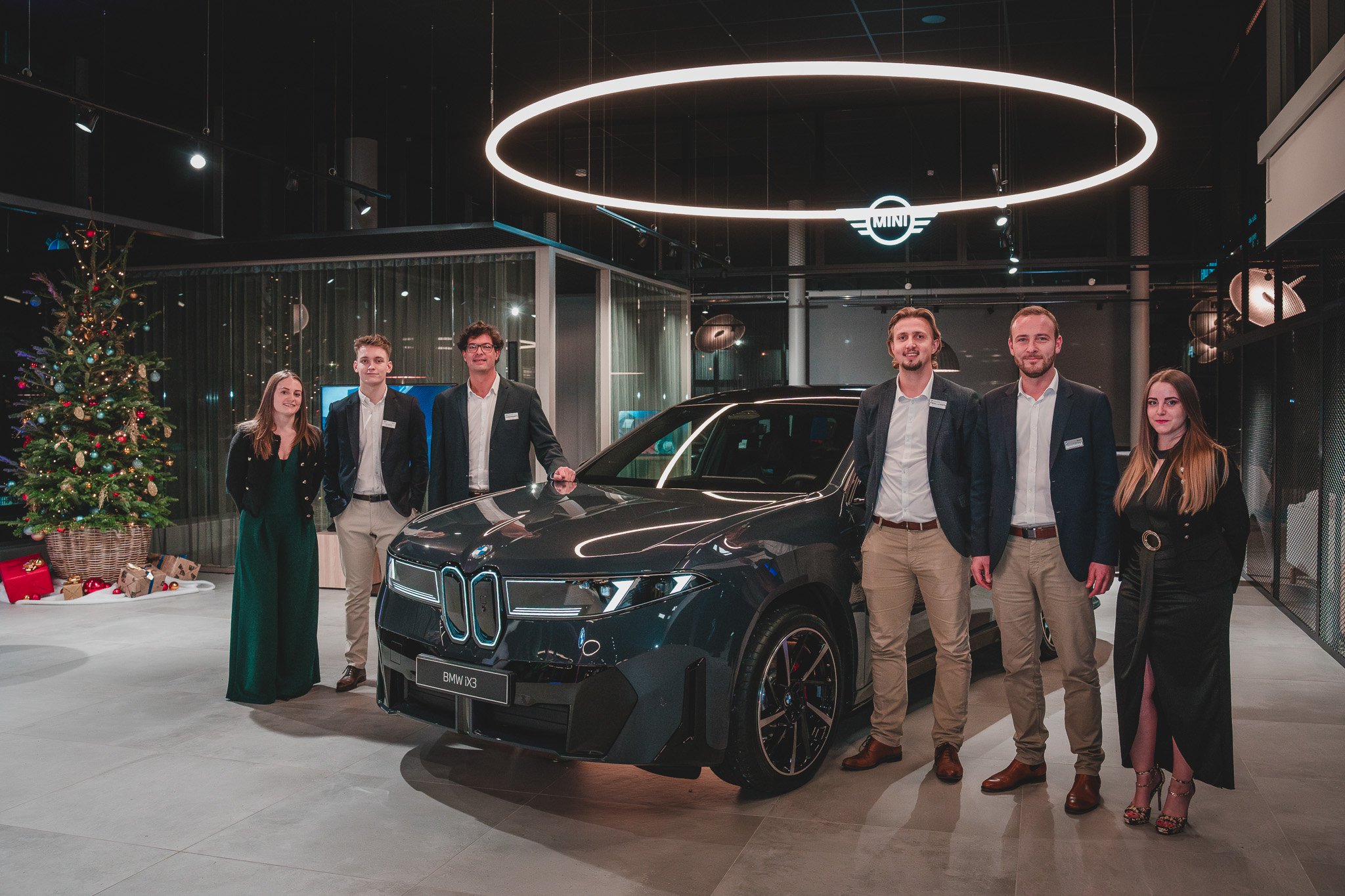 Groupe de six personnes posant avec une voiture électrique BMW iX3 dans un showroom, décoré pour Noël avec un arbre et des cadeaux.