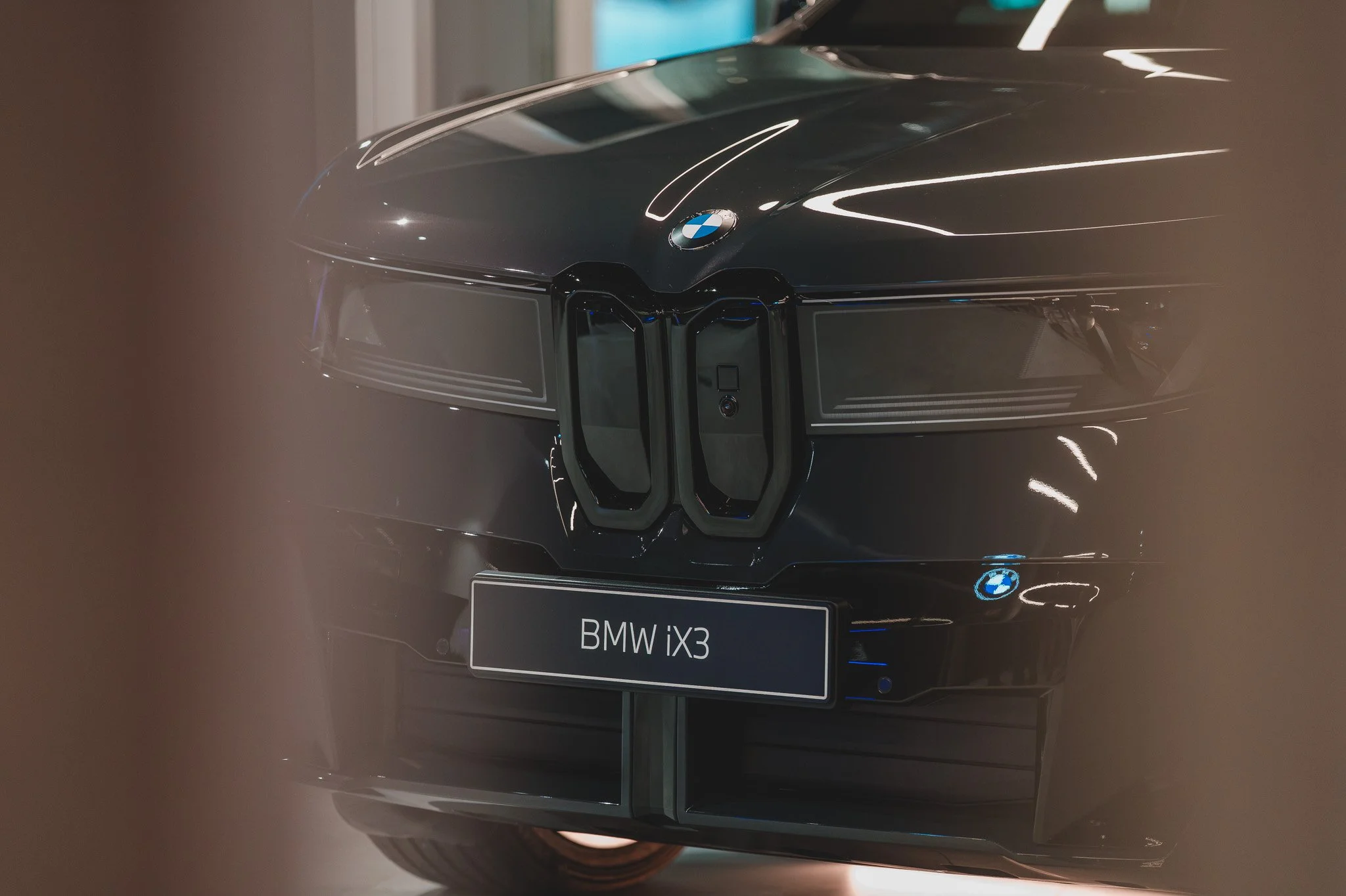 Vue frontale d'une voiture BMW iX3 noire, avec un logo BMW et une plaque d'identification Bavaria, partiellement entourée par un encadrement flou.