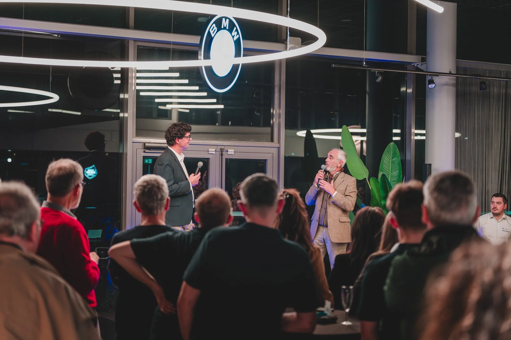 Deux hommes parlant lors d'un événement chez BMW, avec un public en arrière-plan et le logo BMW suspendu au plafond.