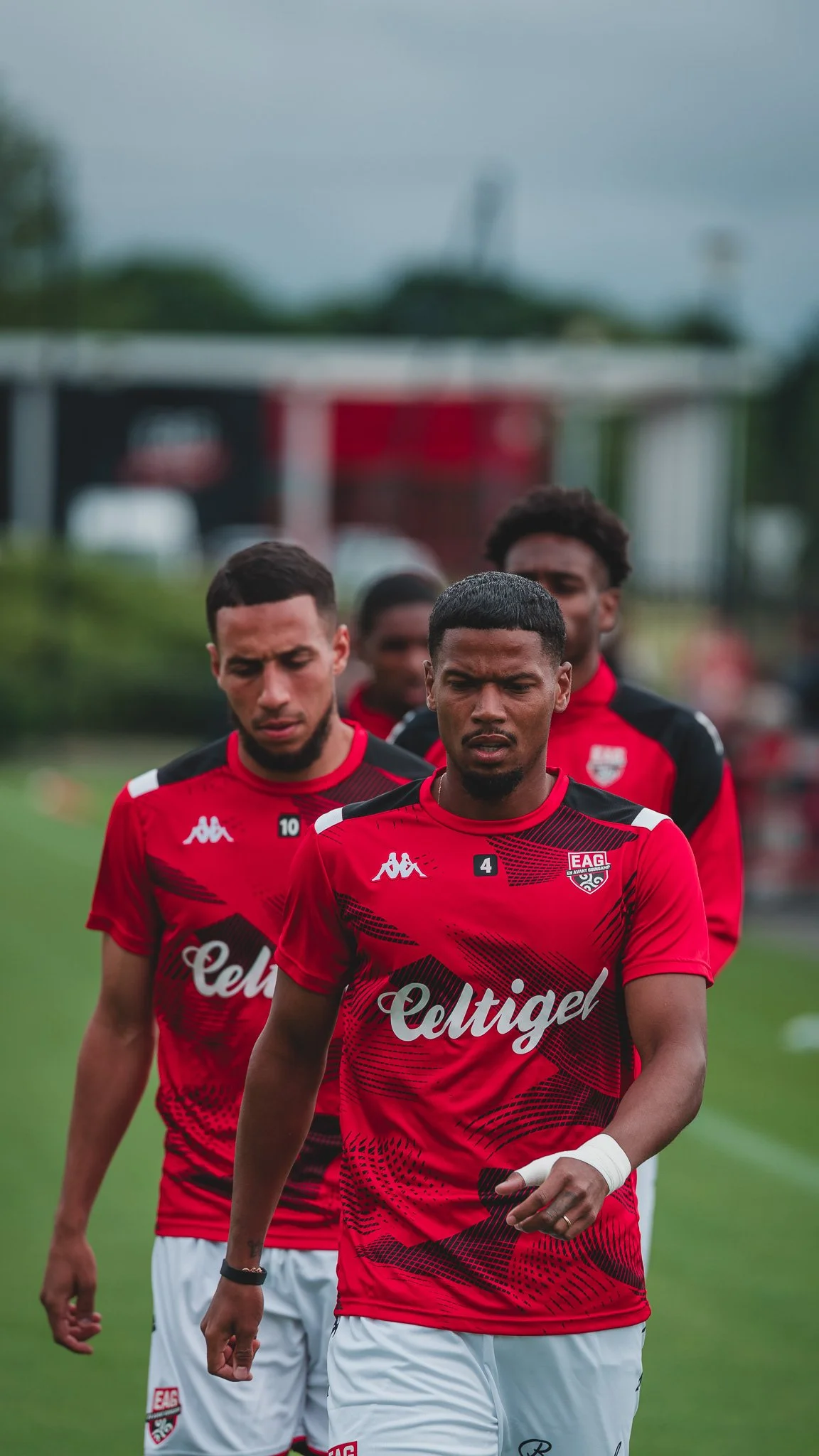 Trois joueurs de football en tenue rouge et blanche, marchant sur un terrain de sport, avec un arrière-plan flou.
