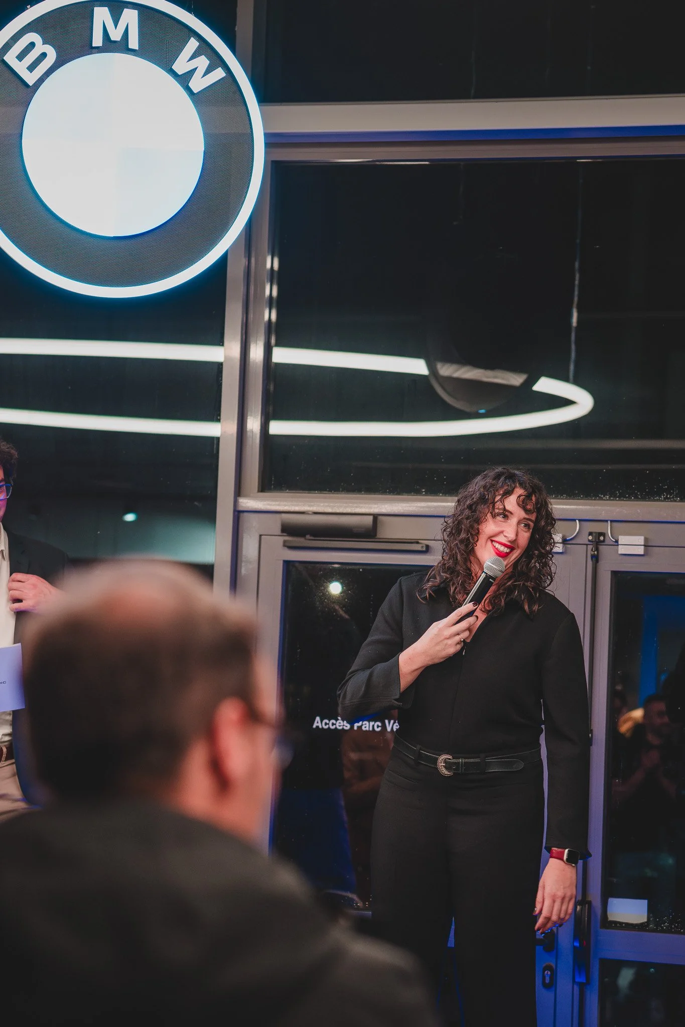 Une femme souriante avec un micro dans une main, portant une tenue noire, lors d'une présentation ou d'un discours dans un lieu intérieur avec un logo BMW en arrière-plan.