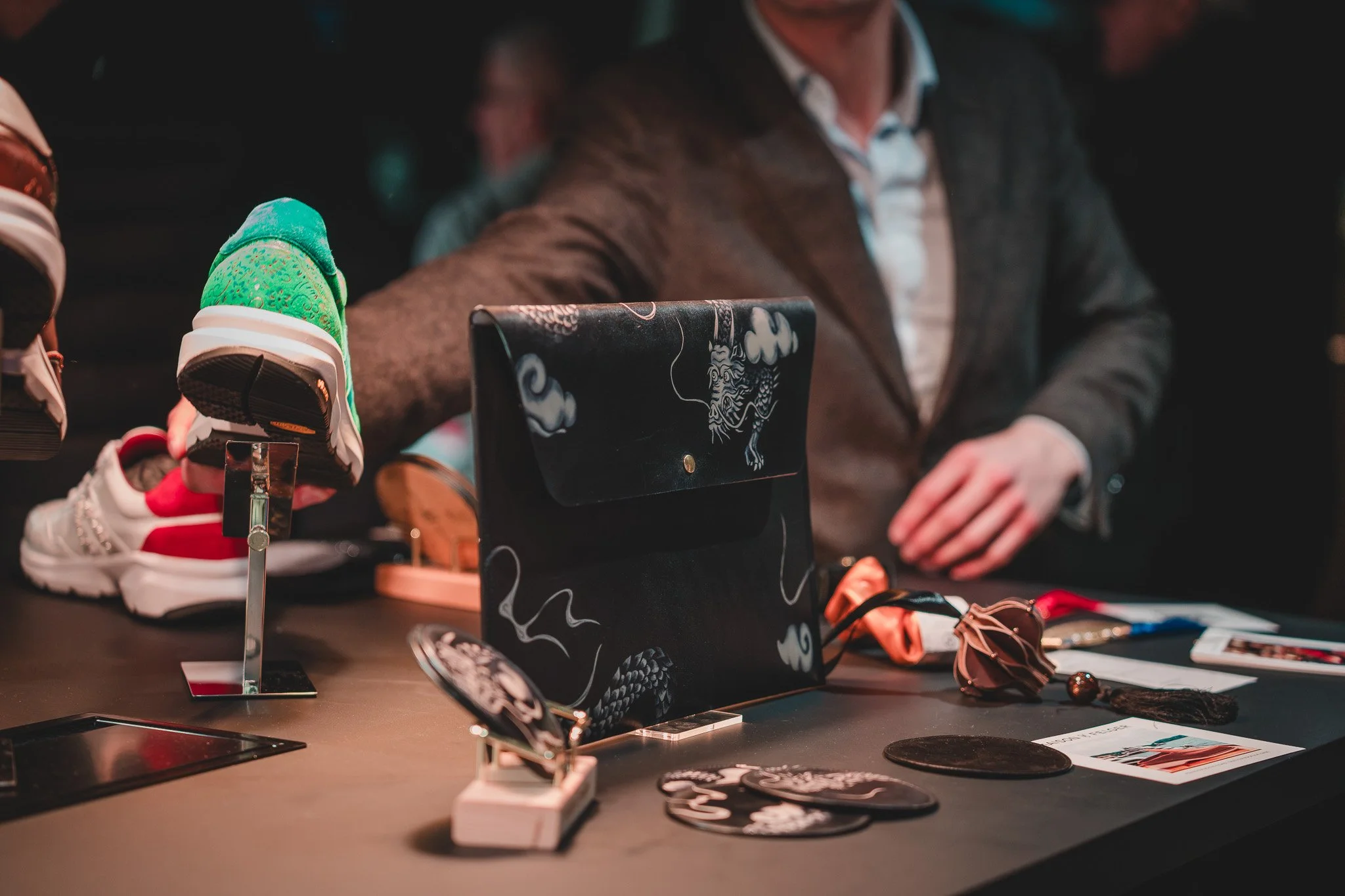 Une table avec des chaussures colorées, un sac à main noir avec un animal dessin, et d'autres accessoires. Un homme en veste est derrière la table.