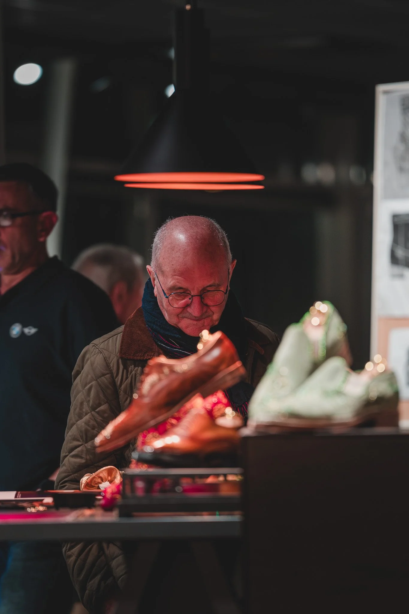 Un homme âgé portant des lunettes, regardant une paire de chaussures de sport dans une boutique. Il y a d'autres personnes en arrière-plan. La scène est éclairée par une lumière rouge au plafond.
