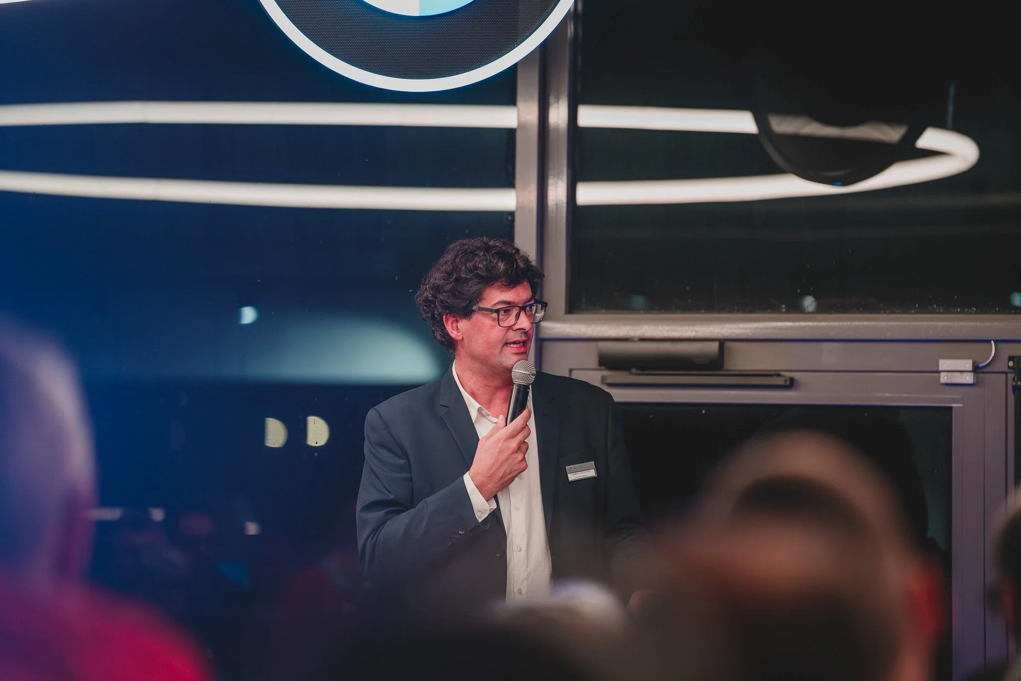 Un homme en costume parle dans un microphone lors d'un événement, avec un fond sombre et un public devant lui.