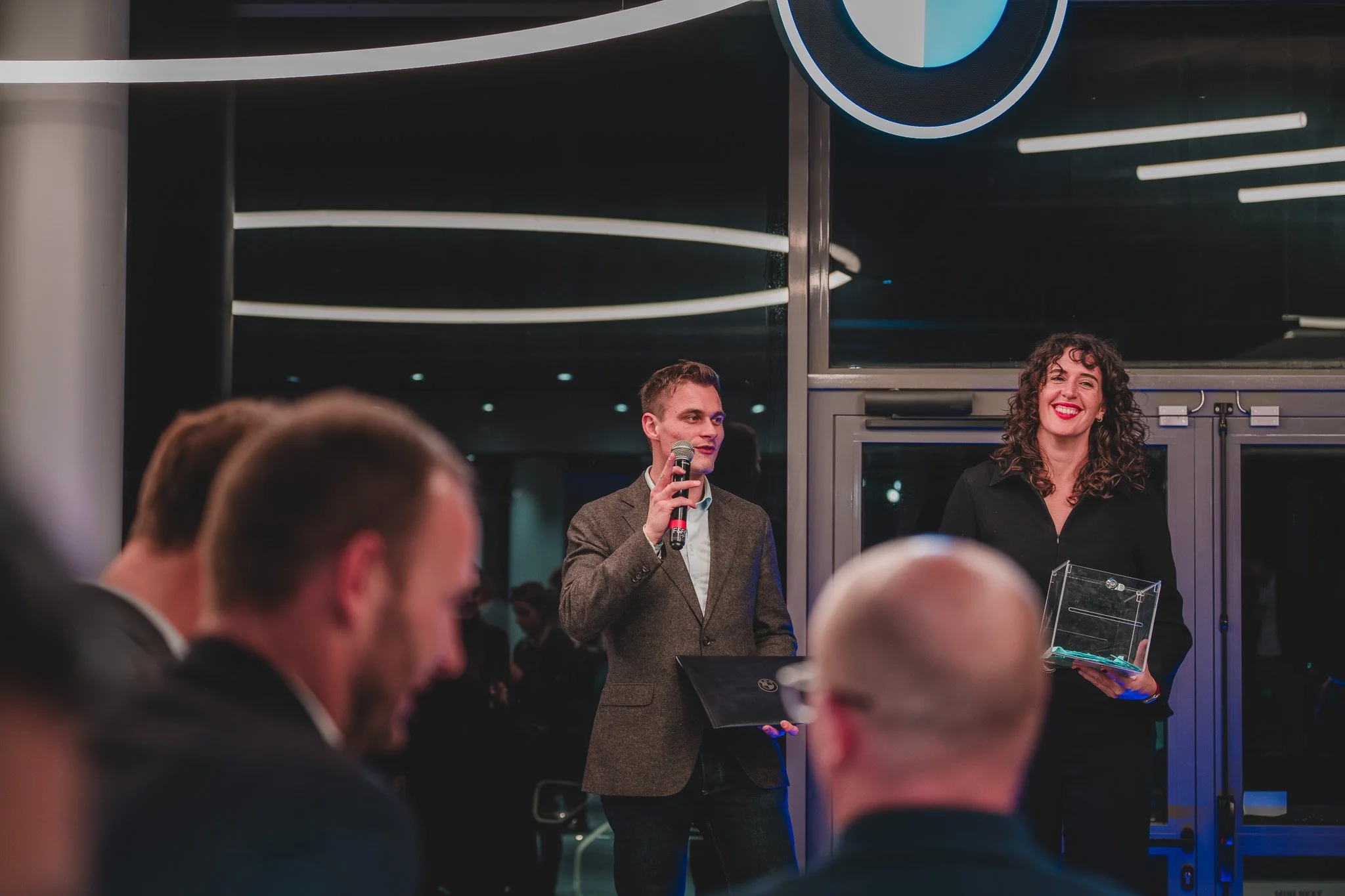 Une cérémonie ou un événement professionnel avec deux personnes debout, une femme tenant un trophée en verre et un homme parlant dans un micro, à l'intérieur, dans une ambiance moderne et élégante.