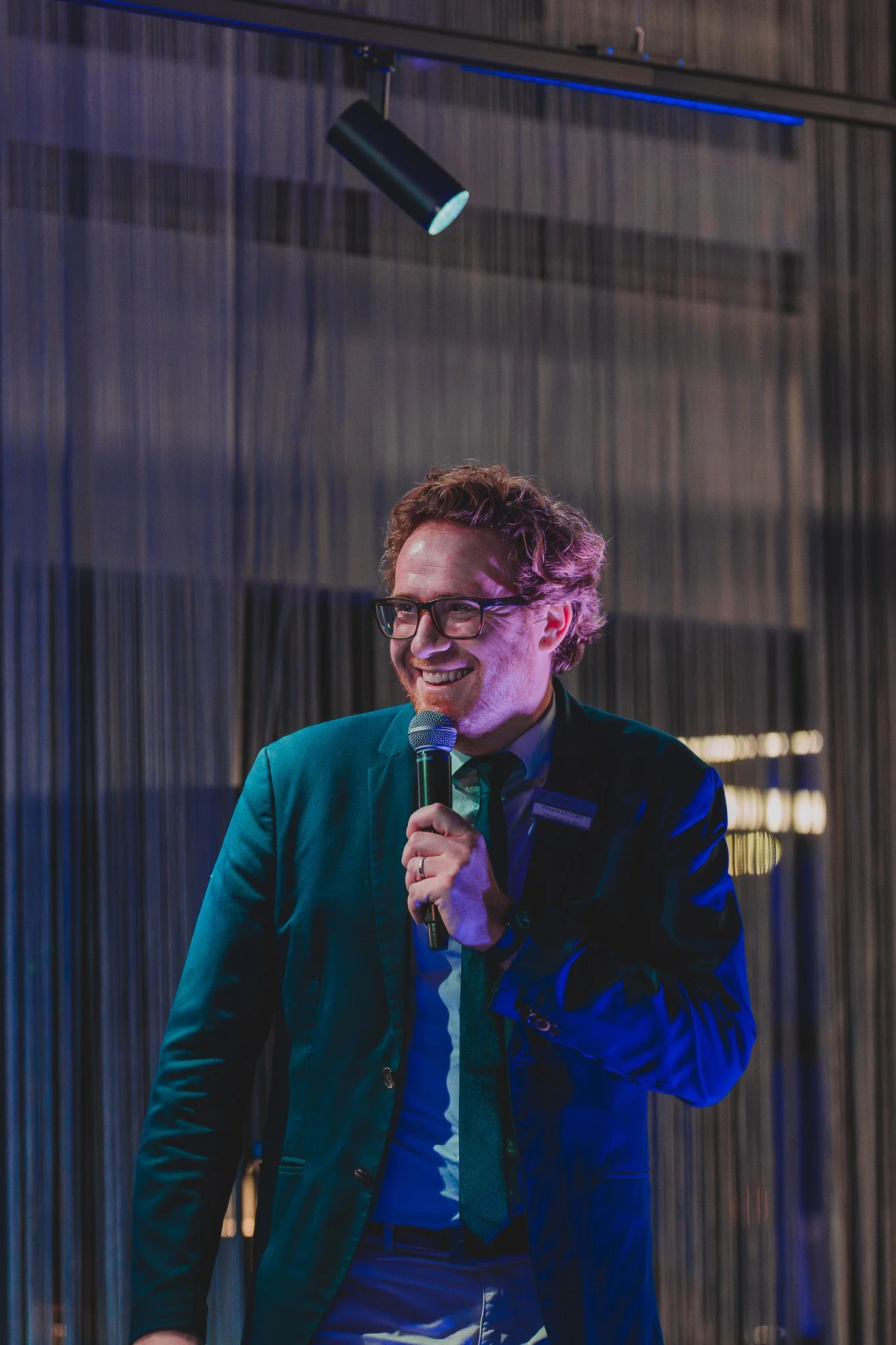 Un homme souriant, portant des lunettes, habillé en costume et cravate, tenant un microphone en train de parler lors d'un événement.