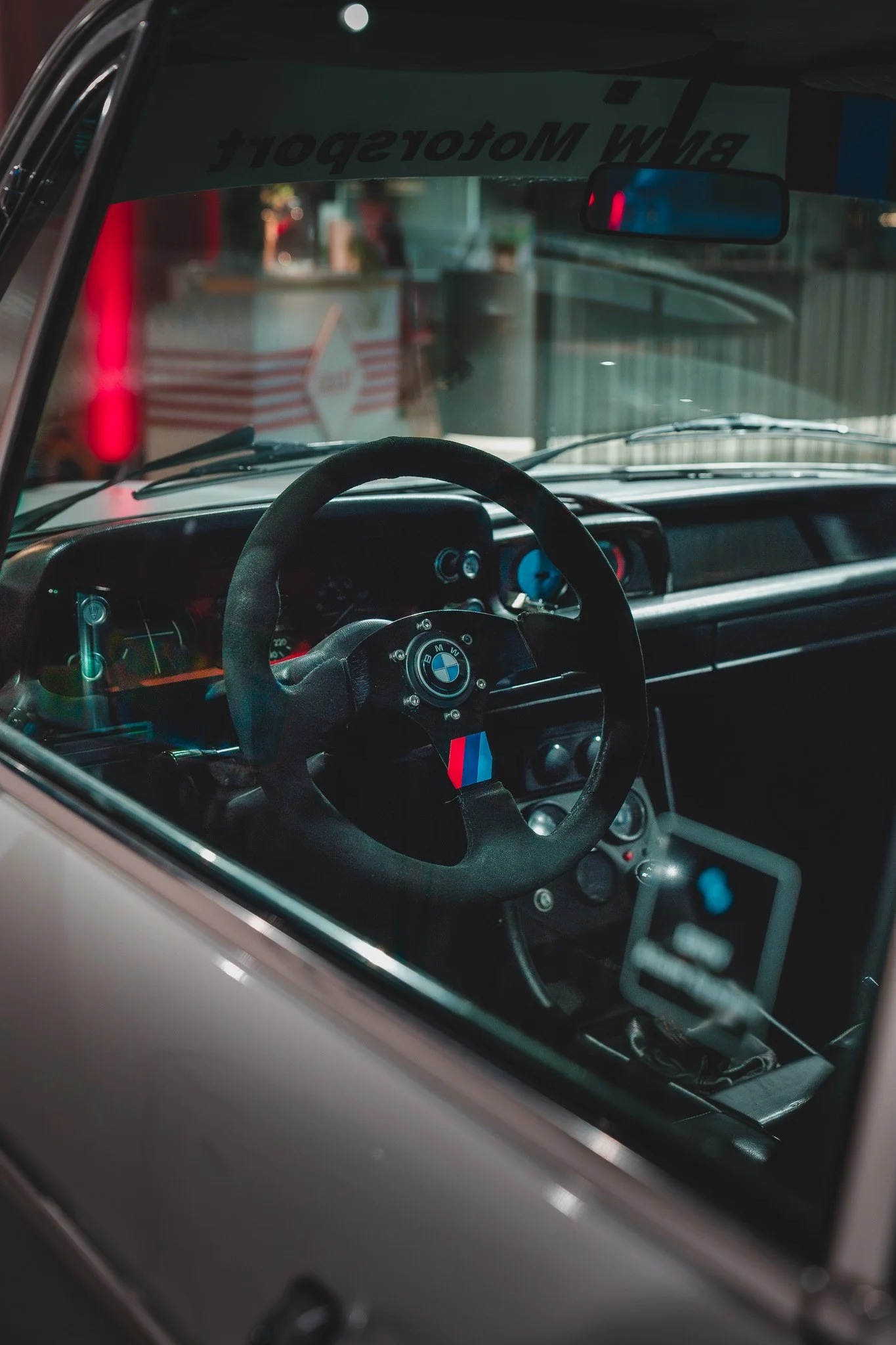 Intérieur d'une voiture de course BMW avec un volant noir, tableau de bord avec plusieurs dials, et un affichage numérique. Le plafond montre un autocollant blanc avec une inscription en anglais. À l'arrière, reflet visible de personnes et d'une stru
