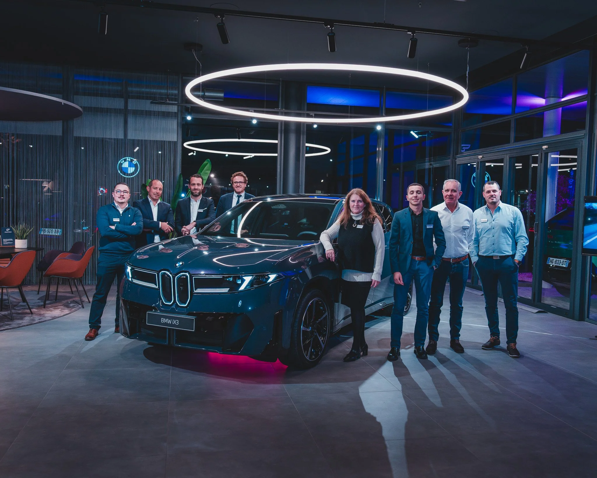Groupe de personnes posant devant une voiture BMW iX3 dans un showroom moderne avec éclairage LED, ambiance nocturne, quelques meubles et décorations