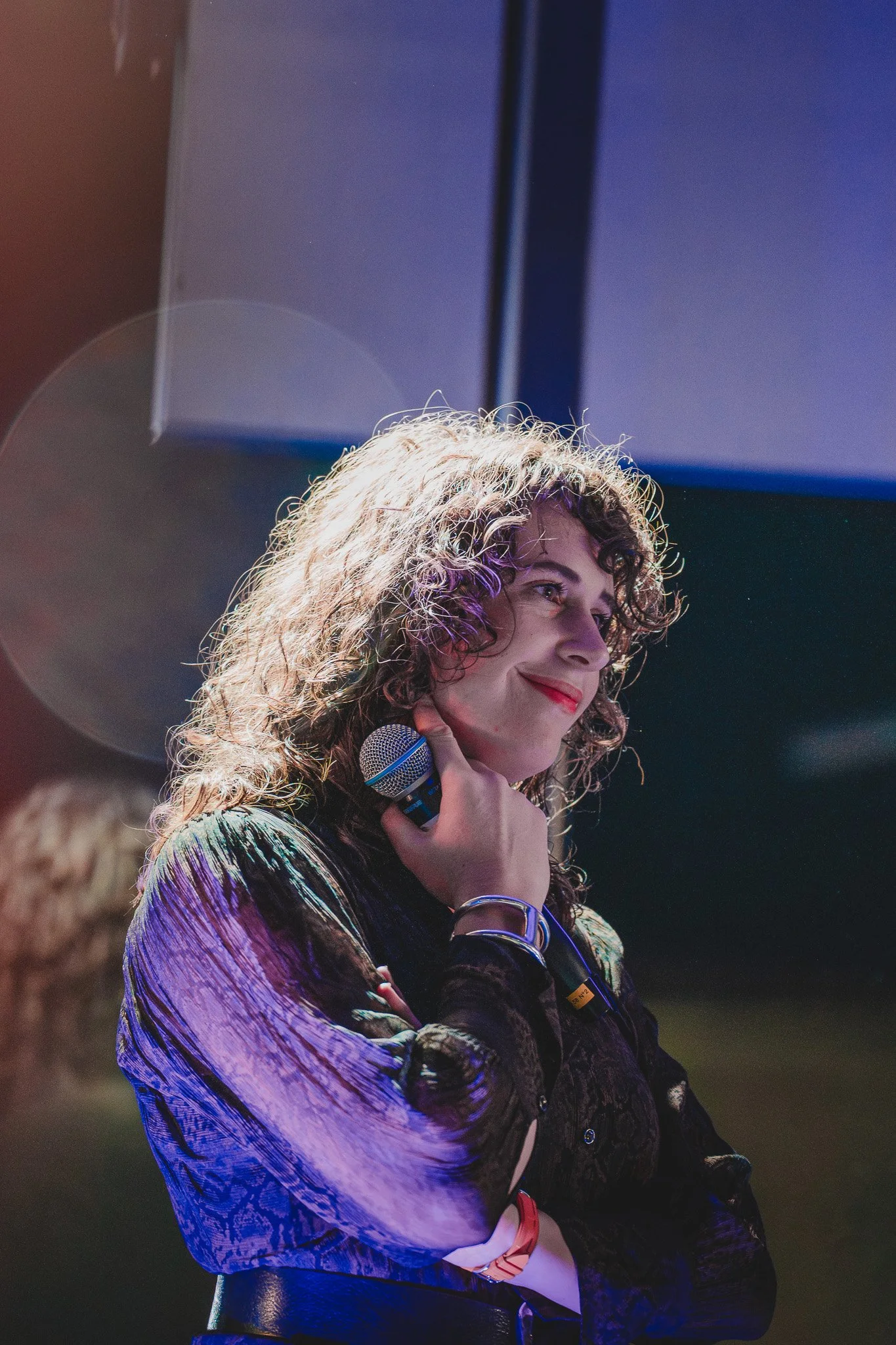 Une femme aux cheveux bouclés, souriante, tenant un micro à la main, en train de participer à un événement ou un spectacle.