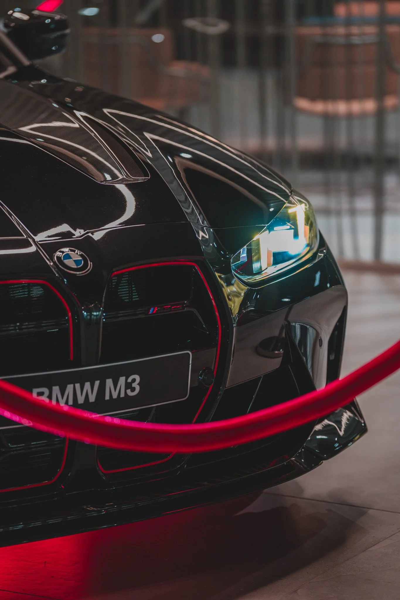 Voiture noire BMW M3 exposée derrière une barrière rouge, mettant en valeur sa carrosserie brillante et ses détails de design moderne.
