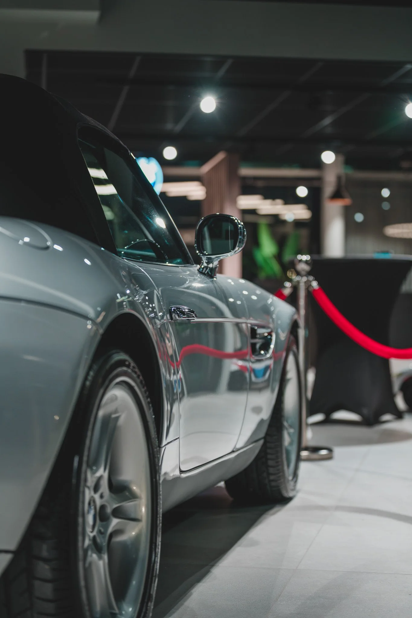 Une voiture de luxe silver chic exposée dans une salle avec un système d'éclairage moderne, protégée par une barrière rouge.