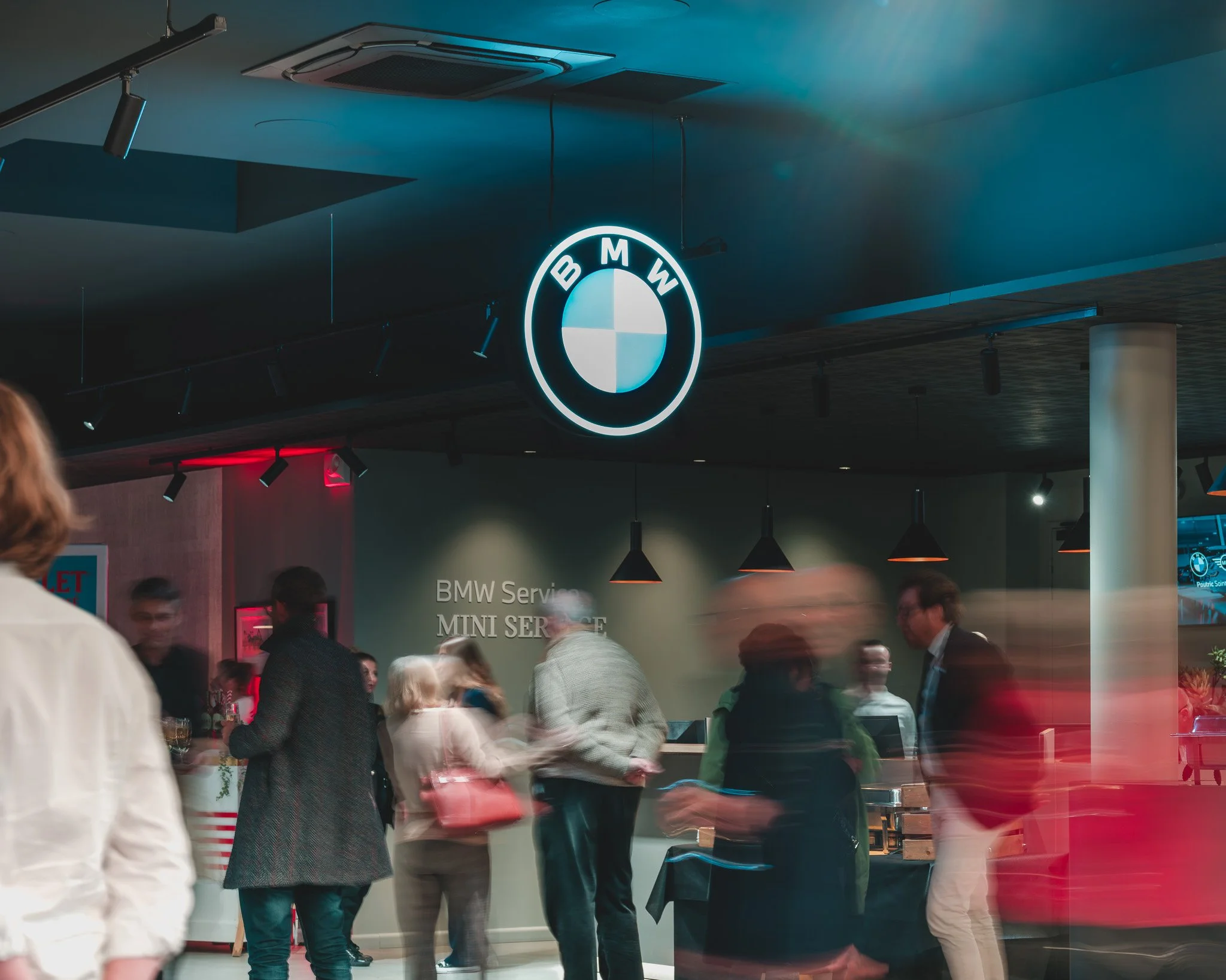 Intérieur d'un centre de service BMW avec un grand logo lumineux au plafond, plusieurs personnes discutant et se déplaçant, ambiance moderne et animée.