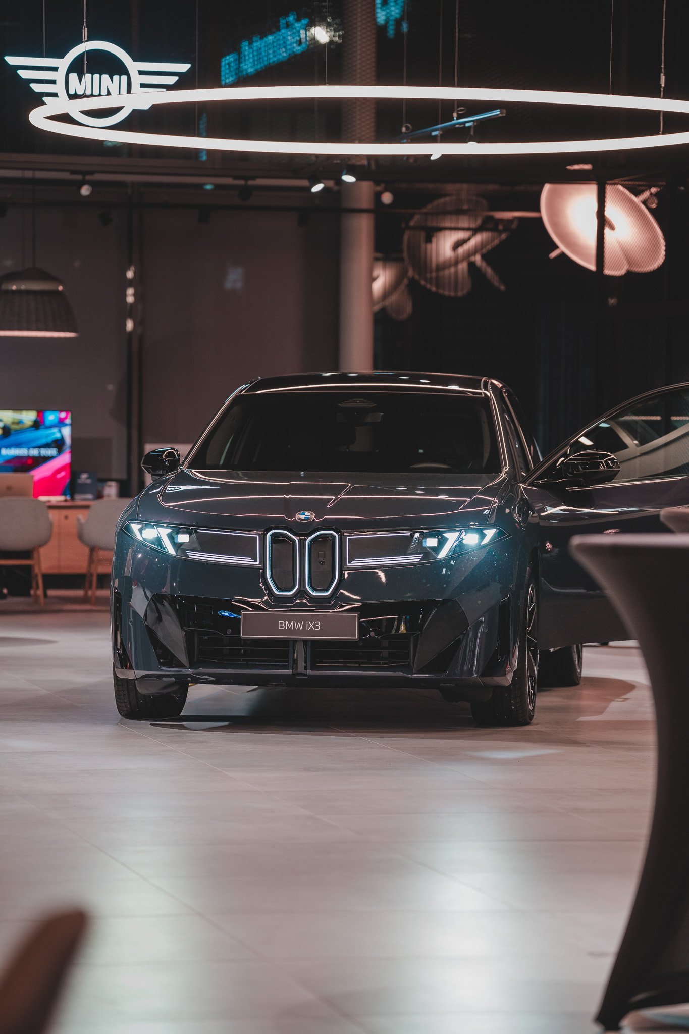Voiture moderne noire BMW iX3 dans un showroom avec éclairage moderne et logo MINI en arrière-plan.