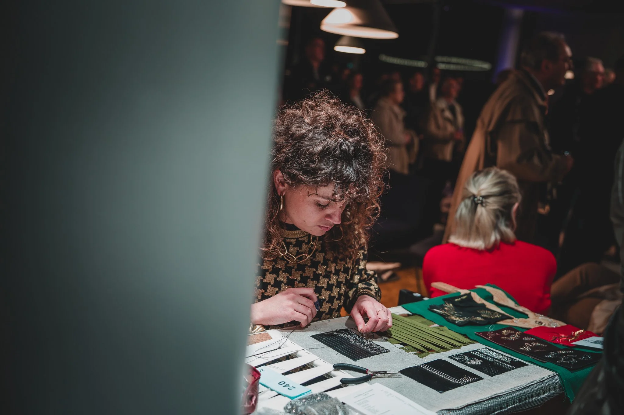 Une femme avec des cheveux bouclés travaillant à une table avec des écharpes ou foulards en tissu de diverses couleurs et motifs, dans un environnement intérieur, apparemment un marché ou une exposition.