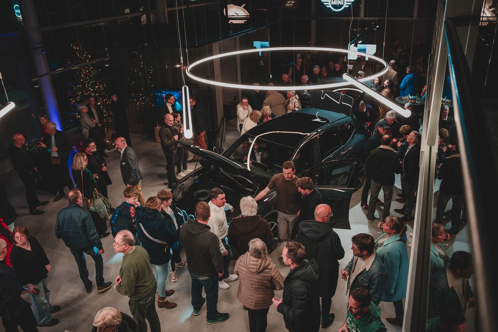 Une foule de personnes à l'intérieur d'un showroom dans la nuit, entourant deux voitures modernes exposées, sous un éclairage décoratif.
