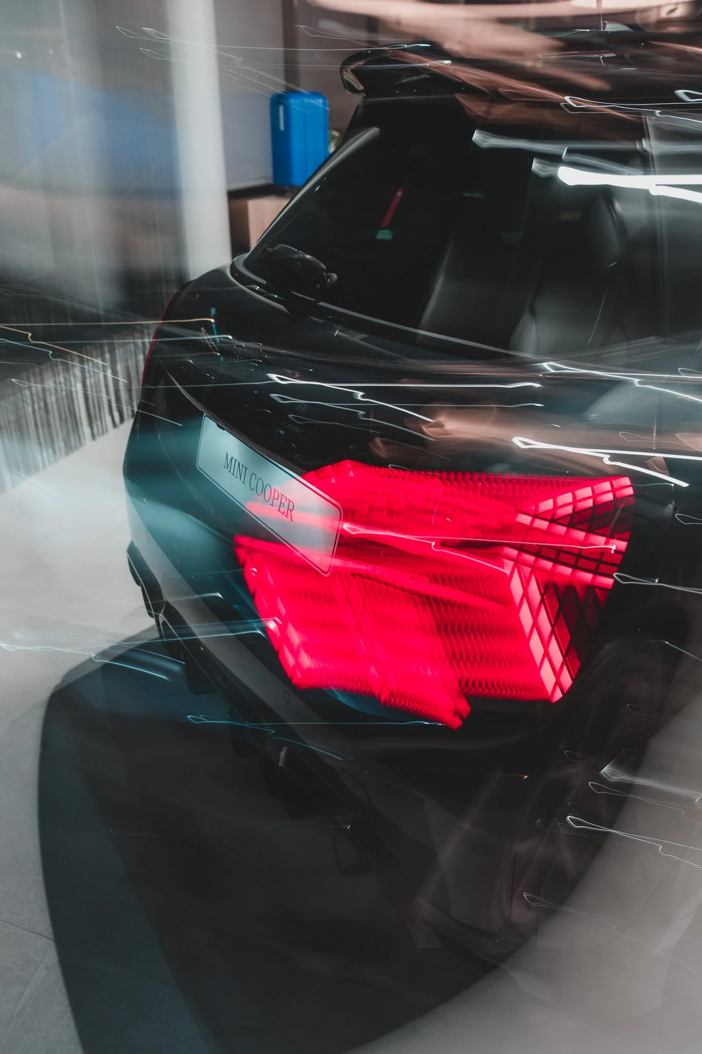 Voiture noire de marque Mini Cooper, avec feux arrière allumés, vue à travers une vitre avec reflets de lumières et un affichage numérique indiquant 'MINI COOPER'.