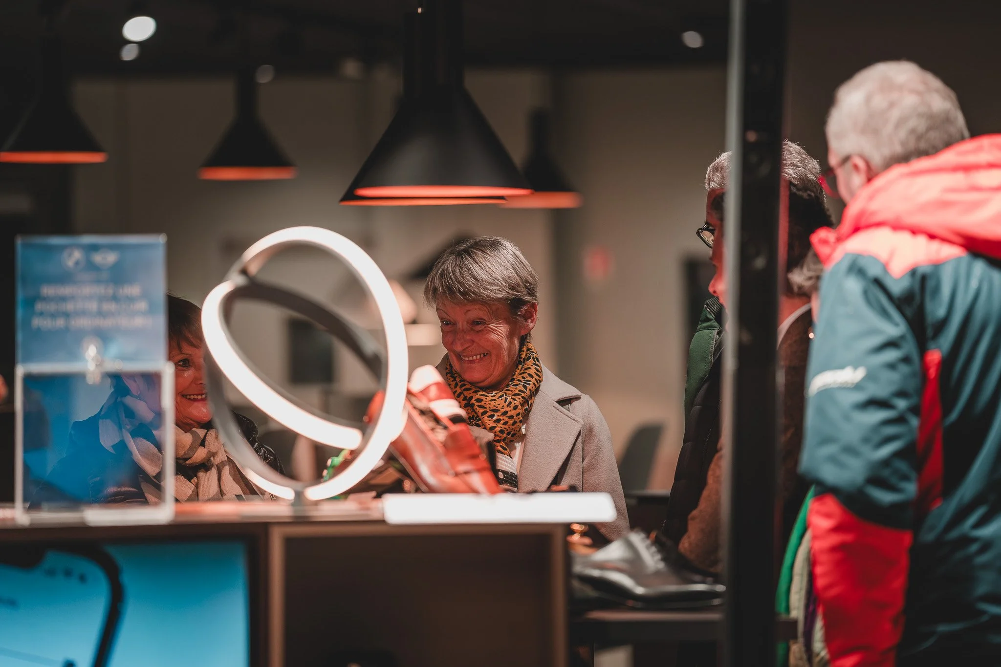 Des personnes souriantes, dont une femme portant un manteau gris et un foulard à motifs, discutent devant un comptoir dans un magasin ou un stand, avec un éclairage chaud et des lampes noires suspendues.