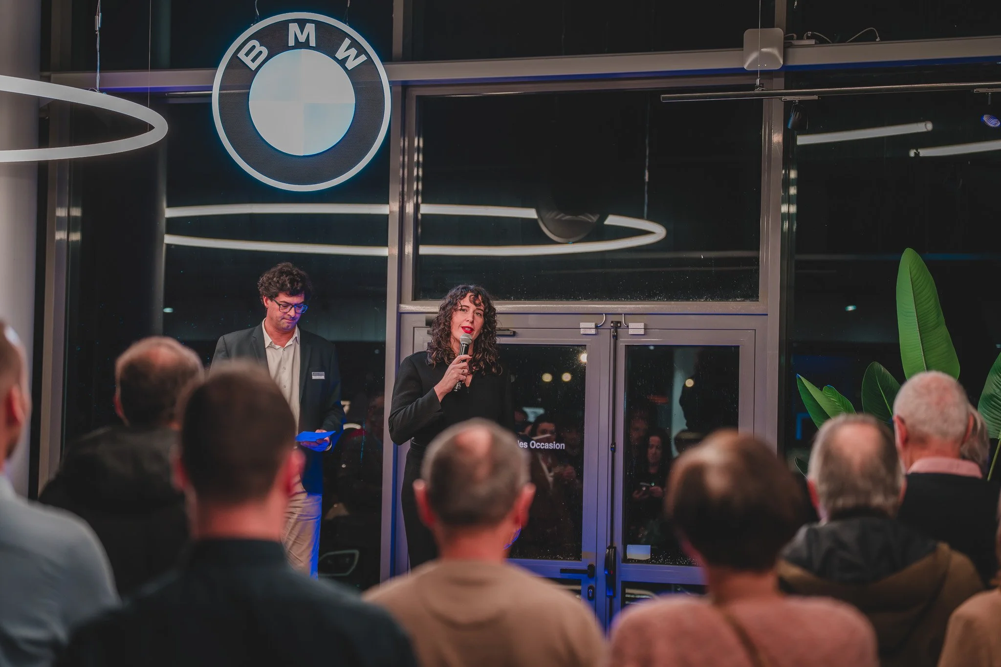 Une femme parle dans un microphone lors d'une conférence ou d'une réunion, avec le logo BMW en arrière-plan, devant un public d'hommes et de femmes.
