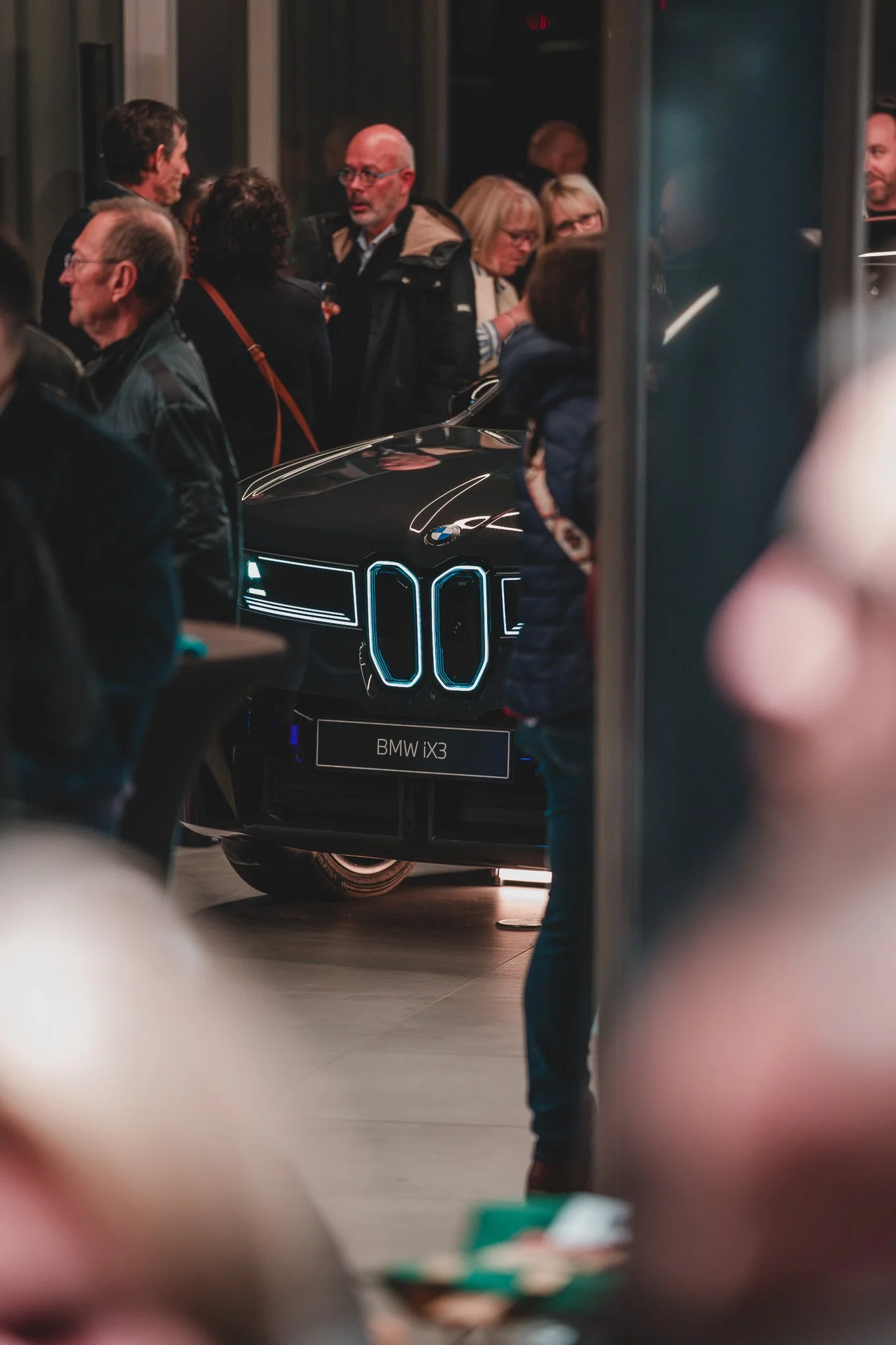 Une foule de personnes autour d'une voiture noire futuriste avec des lignes de lumières bleues, exposée dans une salle, apparemment lors d'un événement de lancement ou d'exposition.