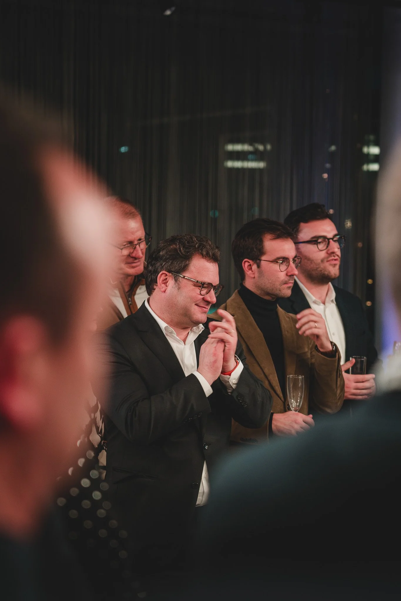 Groupe de quatre hommes en costume lors d'une réception nocturne, certains portent des lunettes, deux tiennent des verres à champagne, un sourit, un autre regarde pensif. Fond sombre avec lumières de ville.