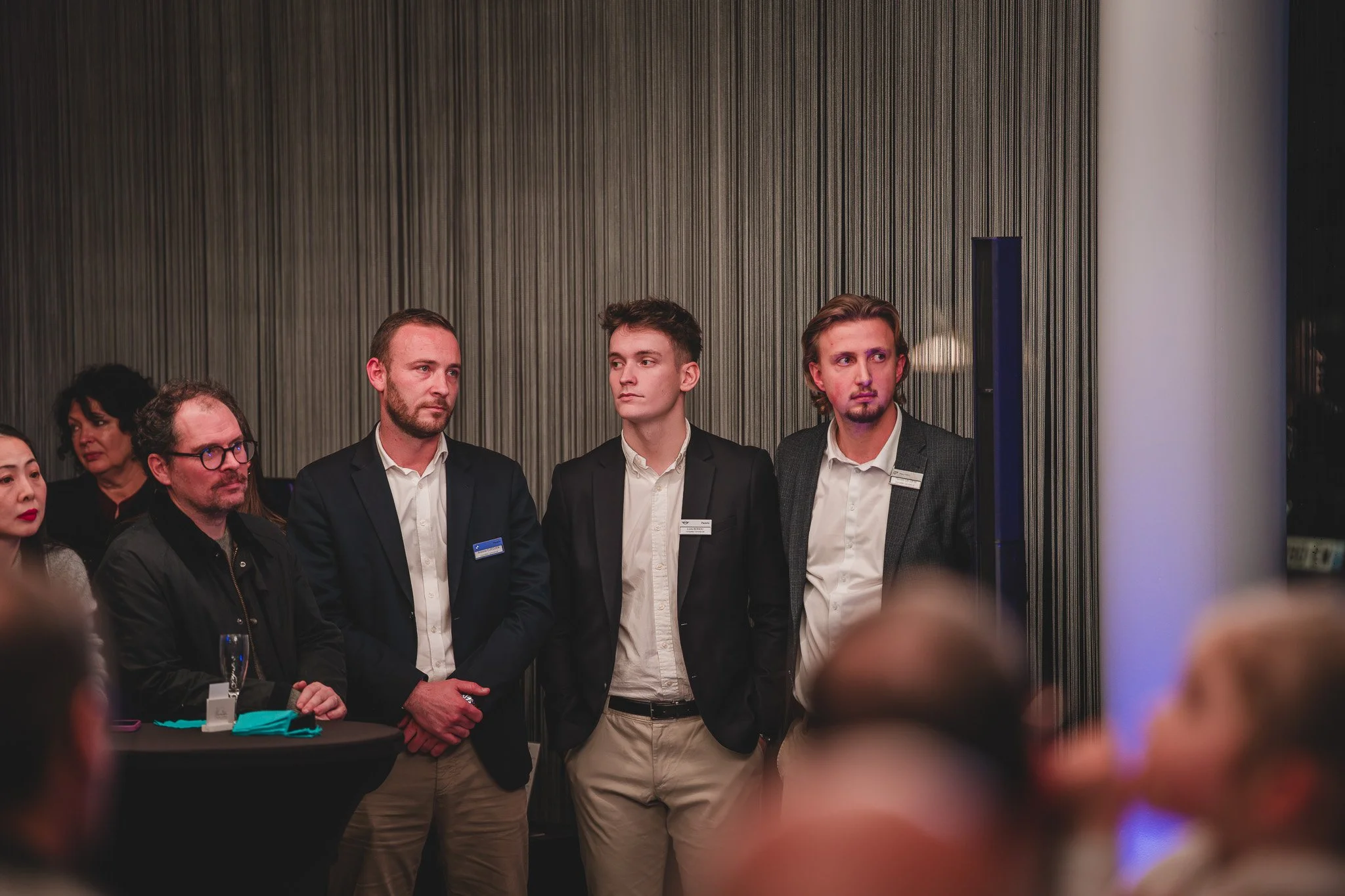 Groupe de jeunes professionnels lors d'un événement, debout contre un mur en arrière-plan.